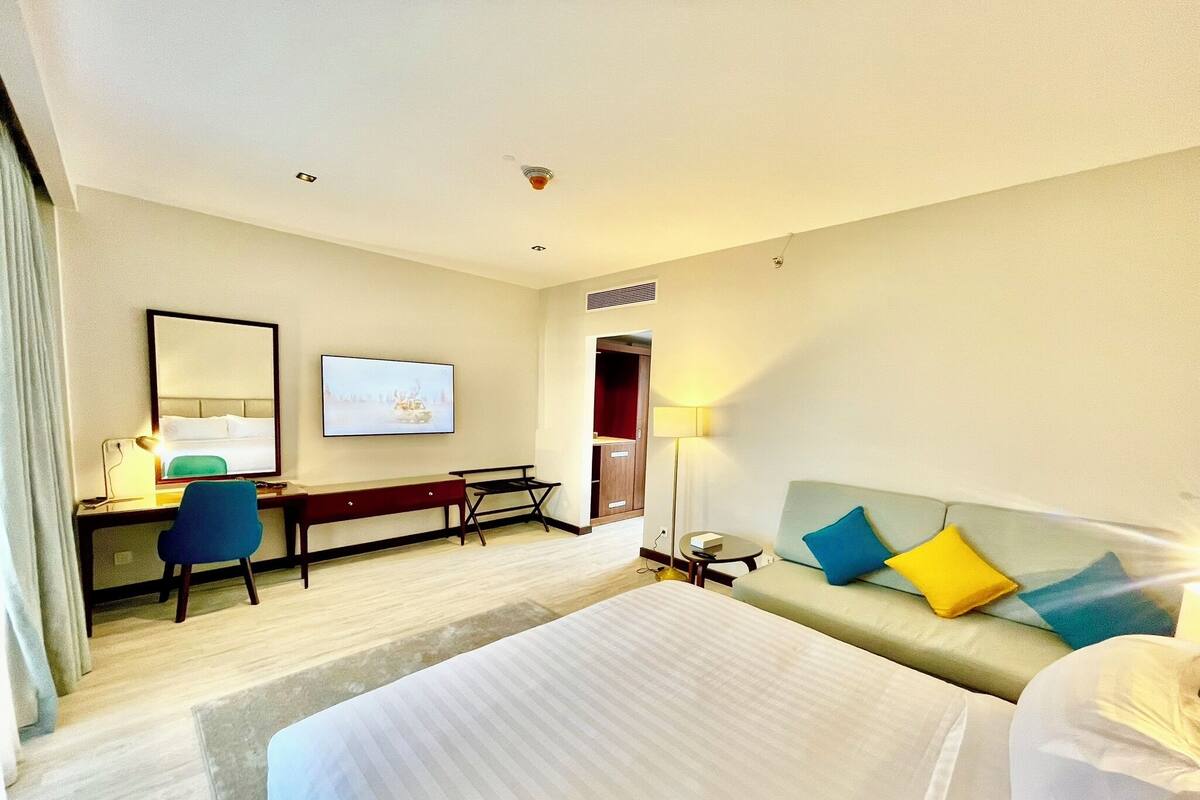 Habitación de hotel con cama, escritorio con silla, sofá con cojines, televisor en la pared y lámpara.