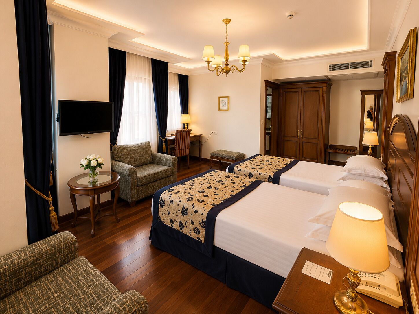 A hotel room with two beds, a desk, a chair, a TV, a chandelier, and a wardrobe.
