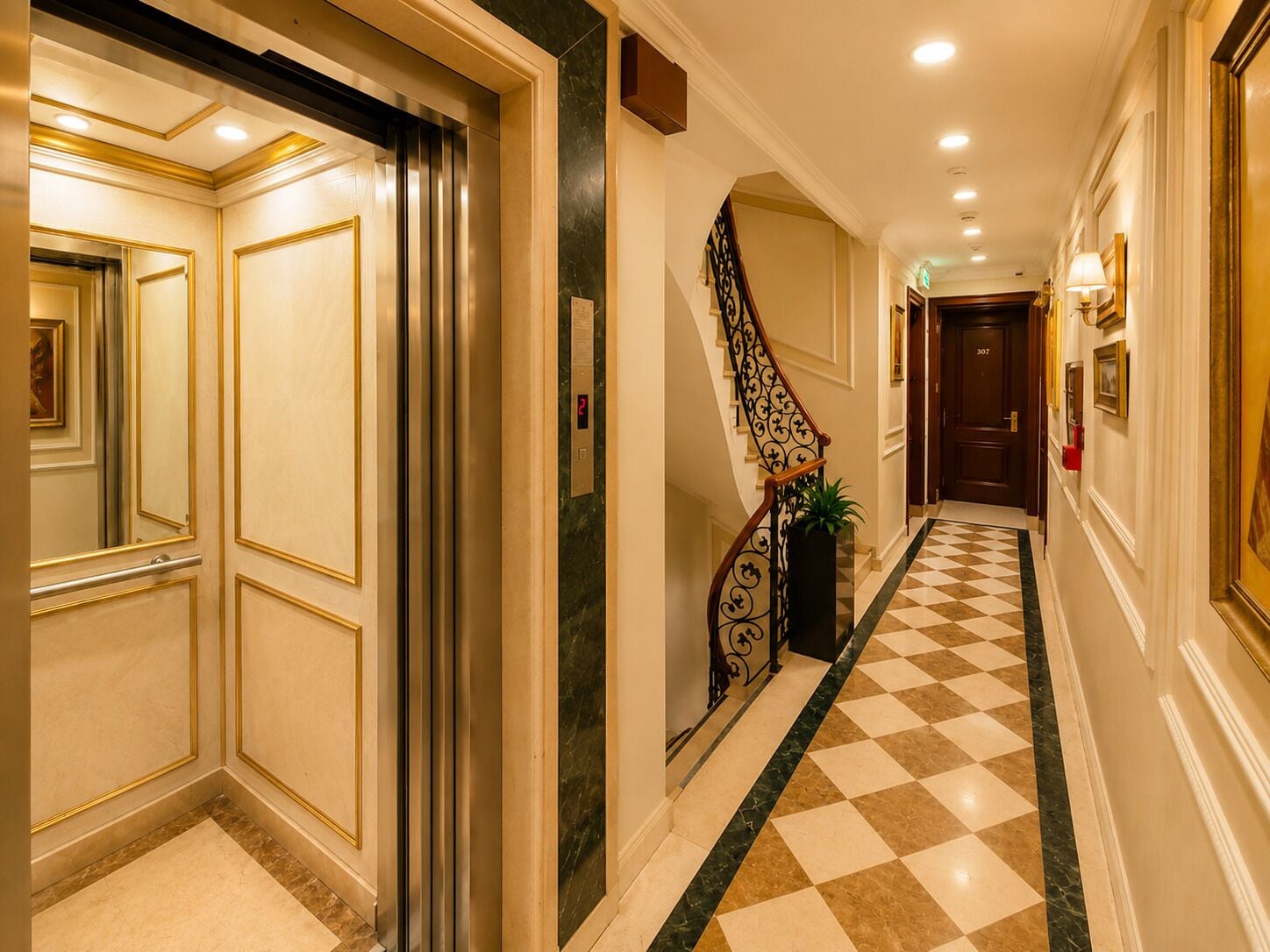 A hotel hallway with a checkered floor, a staircase with a decorative railing, and a door with a digital display.