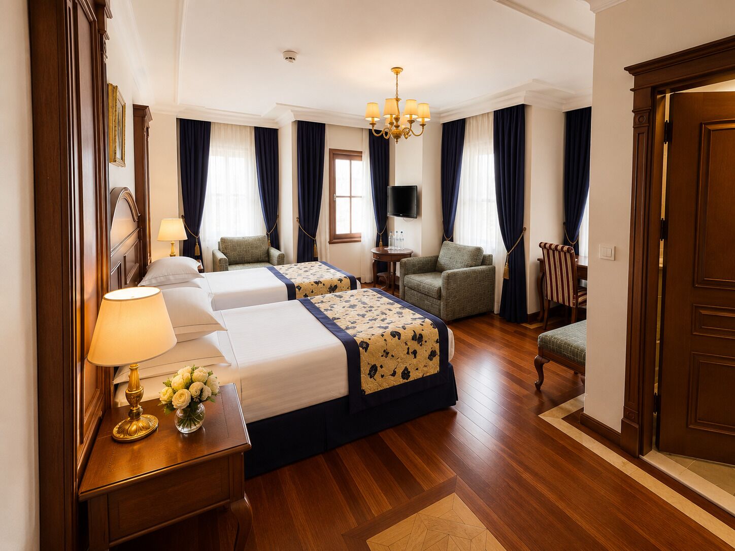A hotel room with two beds, a desk with a lamp, a chair, a television, and a chandelier.