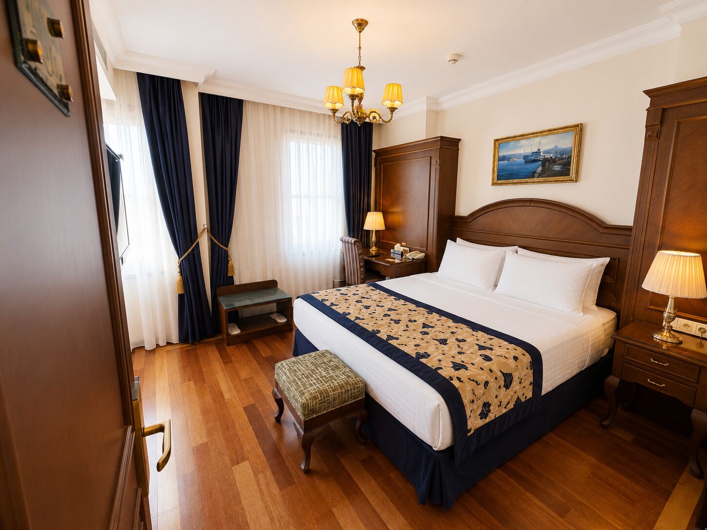 Habitación de hotel con una cama grande, una mesita de noche de madera, un banco, una araña y un cuadro en la pared.
