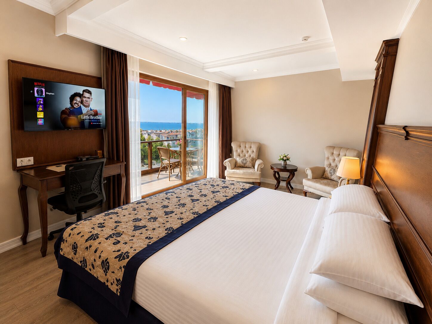 A hotel room with a bed, a desk, a TV, a balcony with a view, and two armchairs.