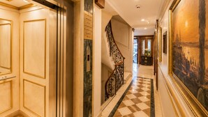 A hotel hallway with a checkered floor, a staircase with ornate railings, and a large framed painting on the wall.