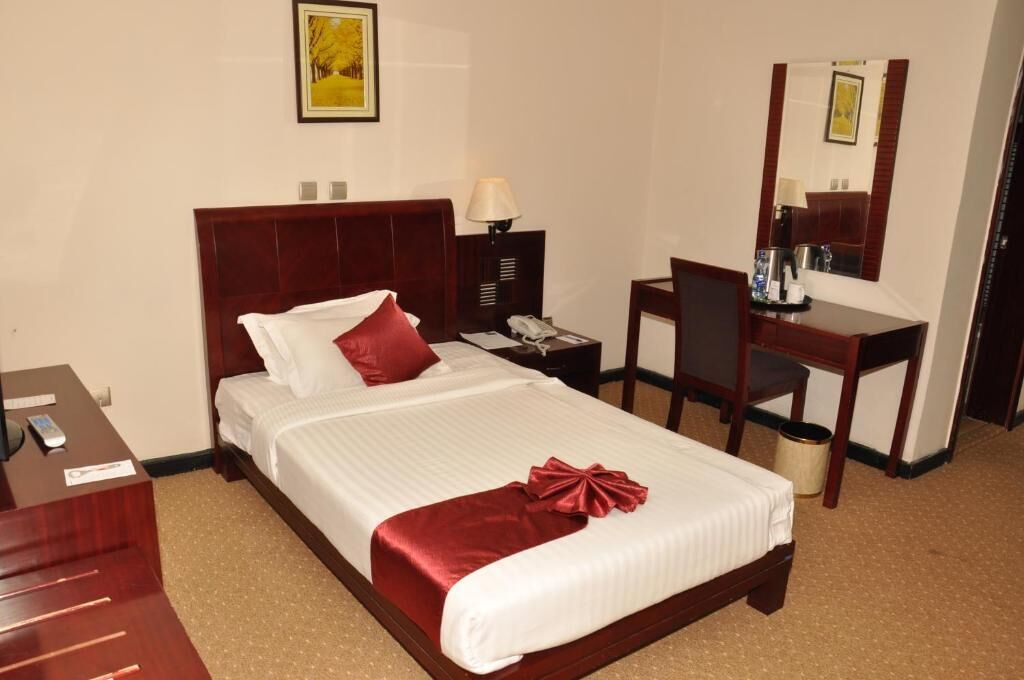 A hotel room with a bed, a desk, a chair, a lamp, a telephone, and a trash can.
