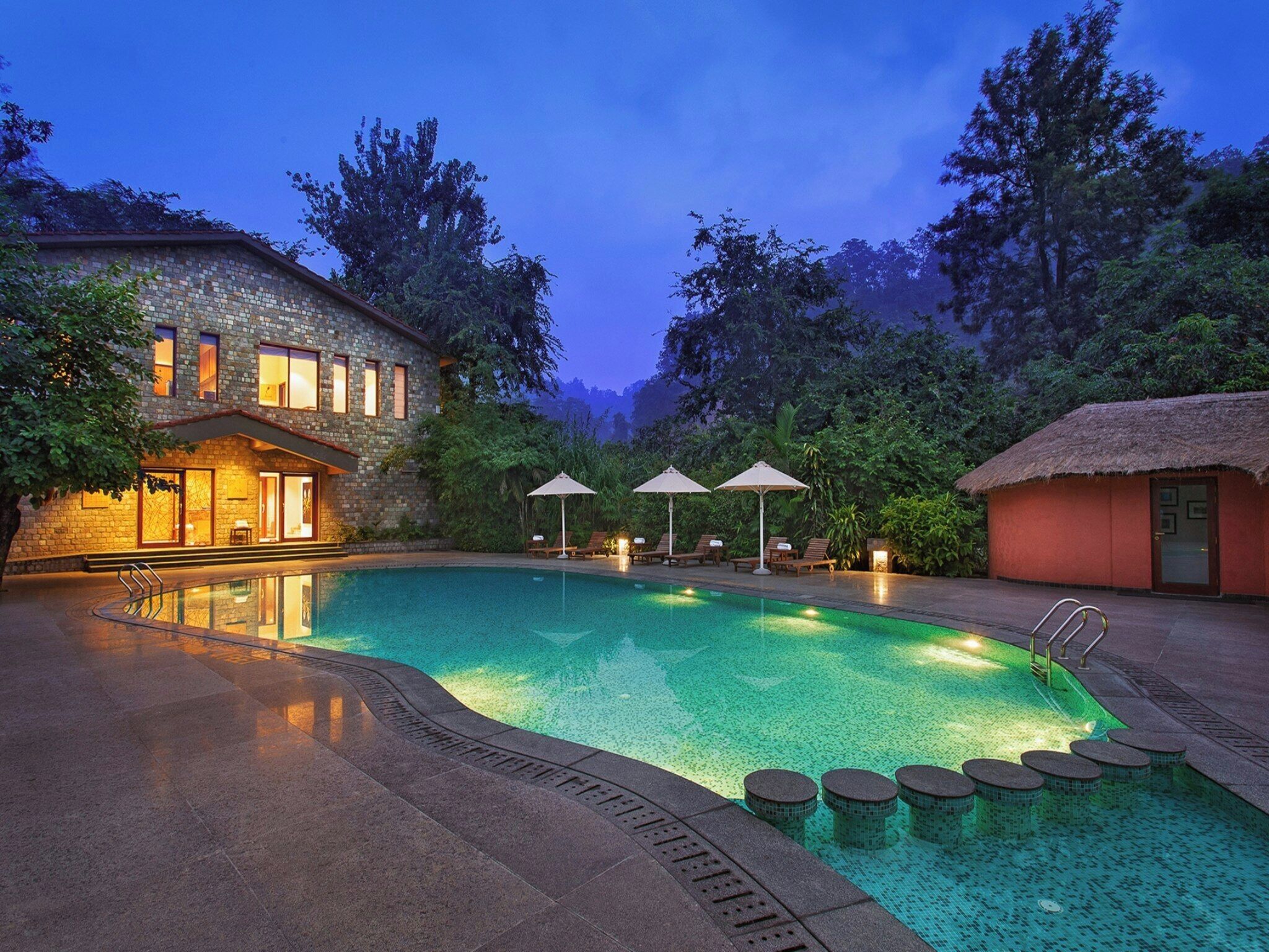 A swimming pool with lounge chairs and umbrellas, surrounded by trees and a stone building.