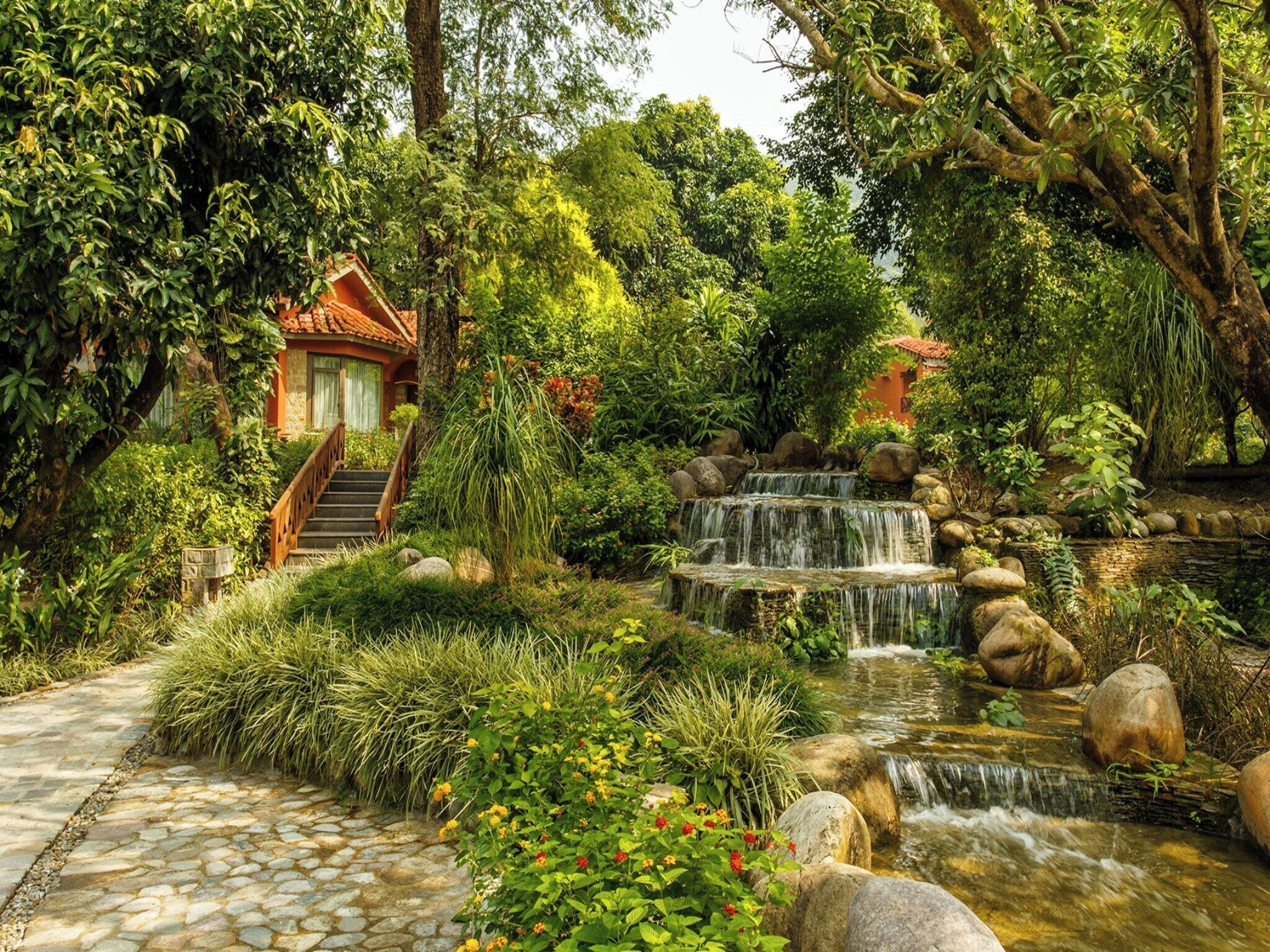 A traditional house with a staircase leading to a tiled roof, surrounded by lush greenery and a small waterfall.
