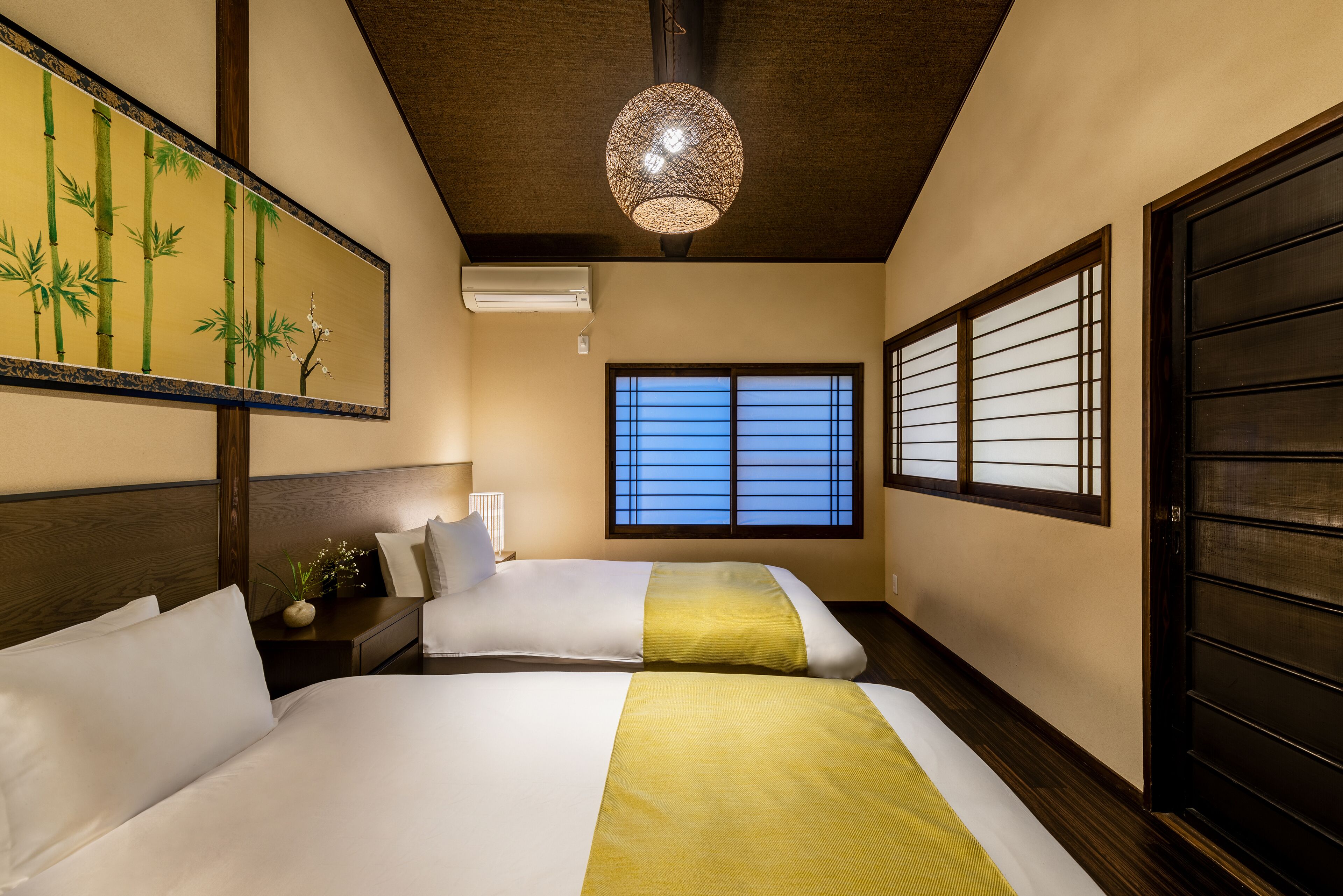 A hotel room with two beds, a large window with shoji screens, a hanging lamp, and a wall-mounted air conditioner.