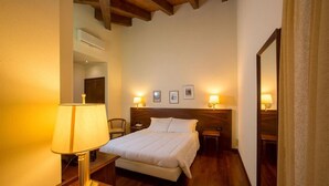 Una habitación de hotel con techo de madera, una cama con ropa de cama blanca, dos mesitas de noche con lámparas, una mesita auxiliar de madera y un espejo en la pared.