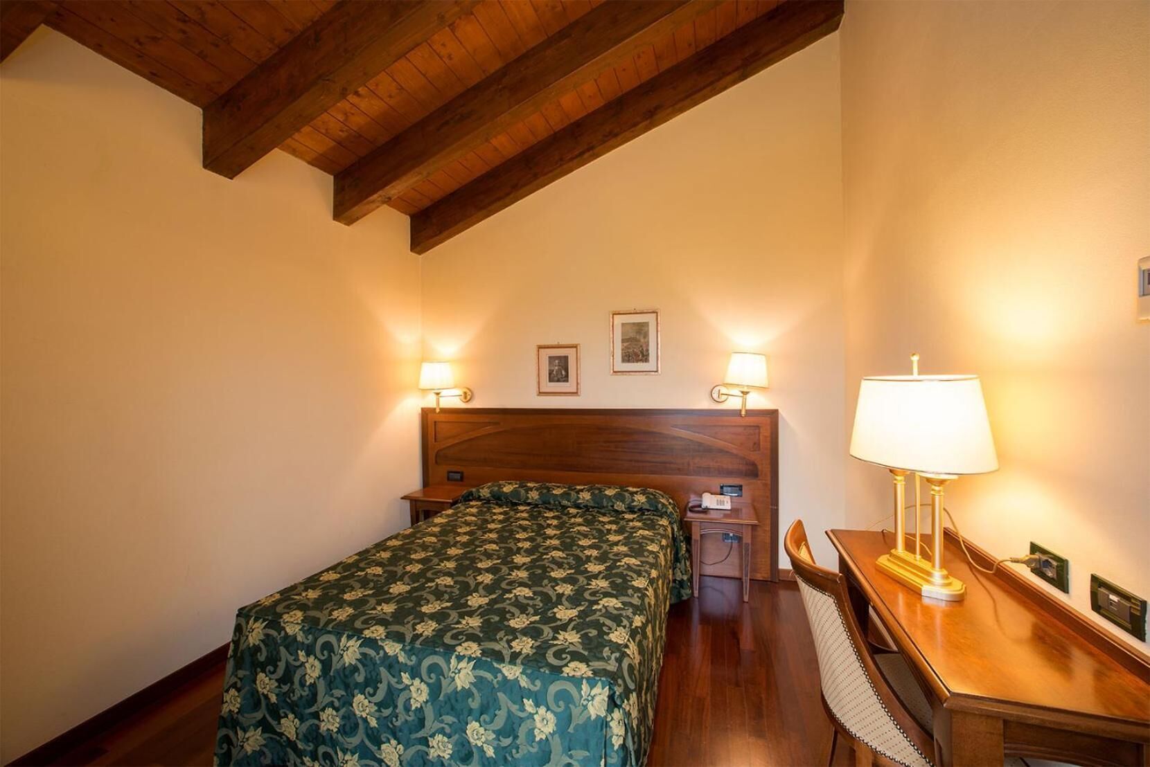 Una camera d'albergo con soffitto in legno, un letto con copripiumino floreale, due abat-jour, una scrivania con lampada e una sedia.