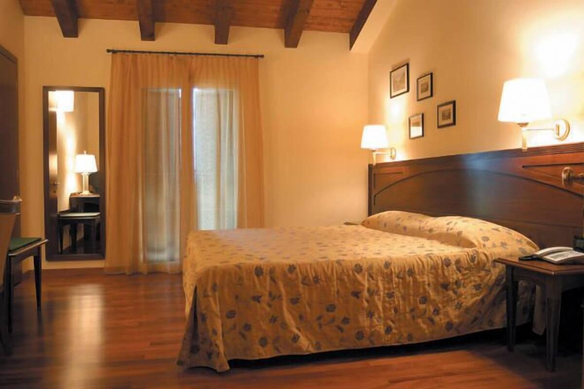 Ein Schlafzimmer mit einem hölzernen Bett, einem Nachttisch, einem Sessel, einem Schreibtisch, einem Spiegel und einem Fenster mit Vorhängen.