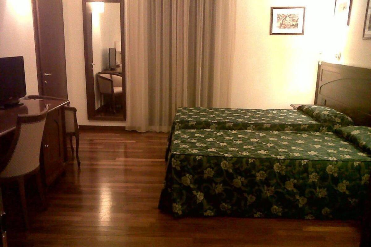 Ein Hotelzimmer mit Holzboden, einem Schreibtisch mit Stuhl, zwei Betten mit Blumenmuster-Bettwäsche, einem Spiegel und einem gerahmten Bild an der Wand.
