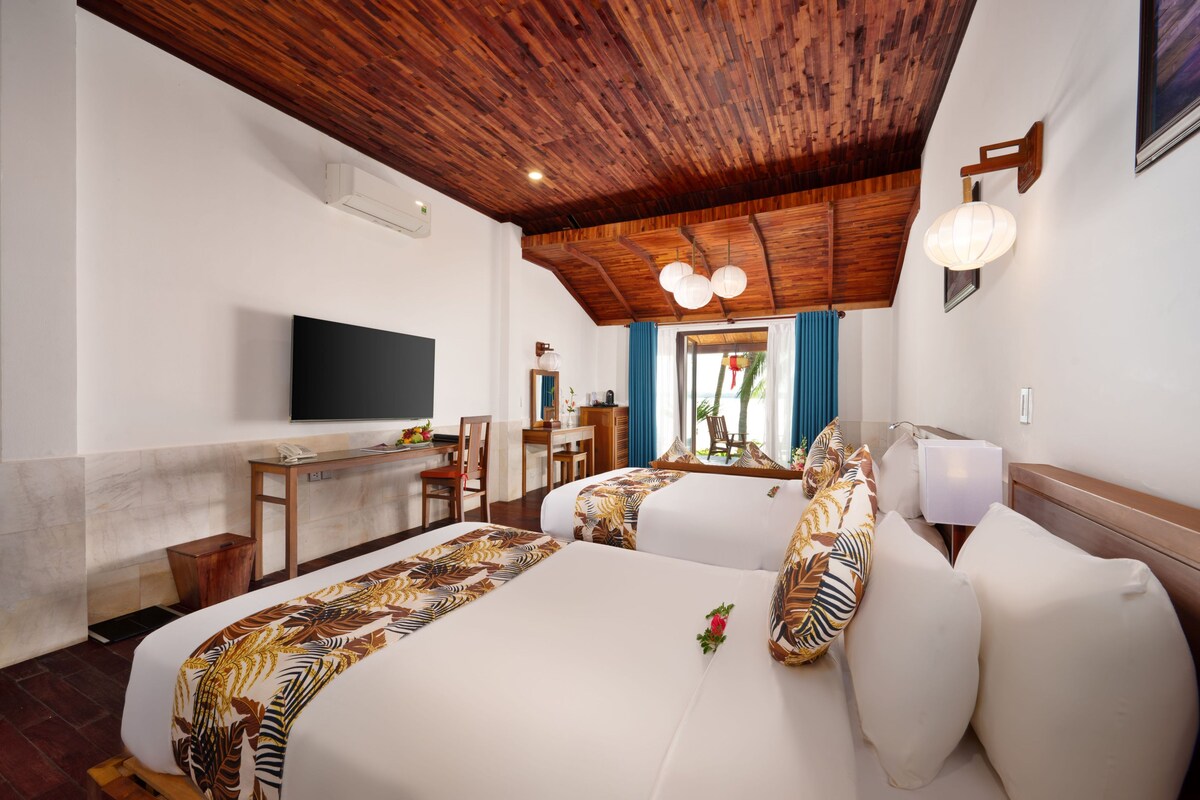 A hotel room with a wooden ceiling, a flat-screen TV, a desk with a chair, and two beds with patterned bedspreads.