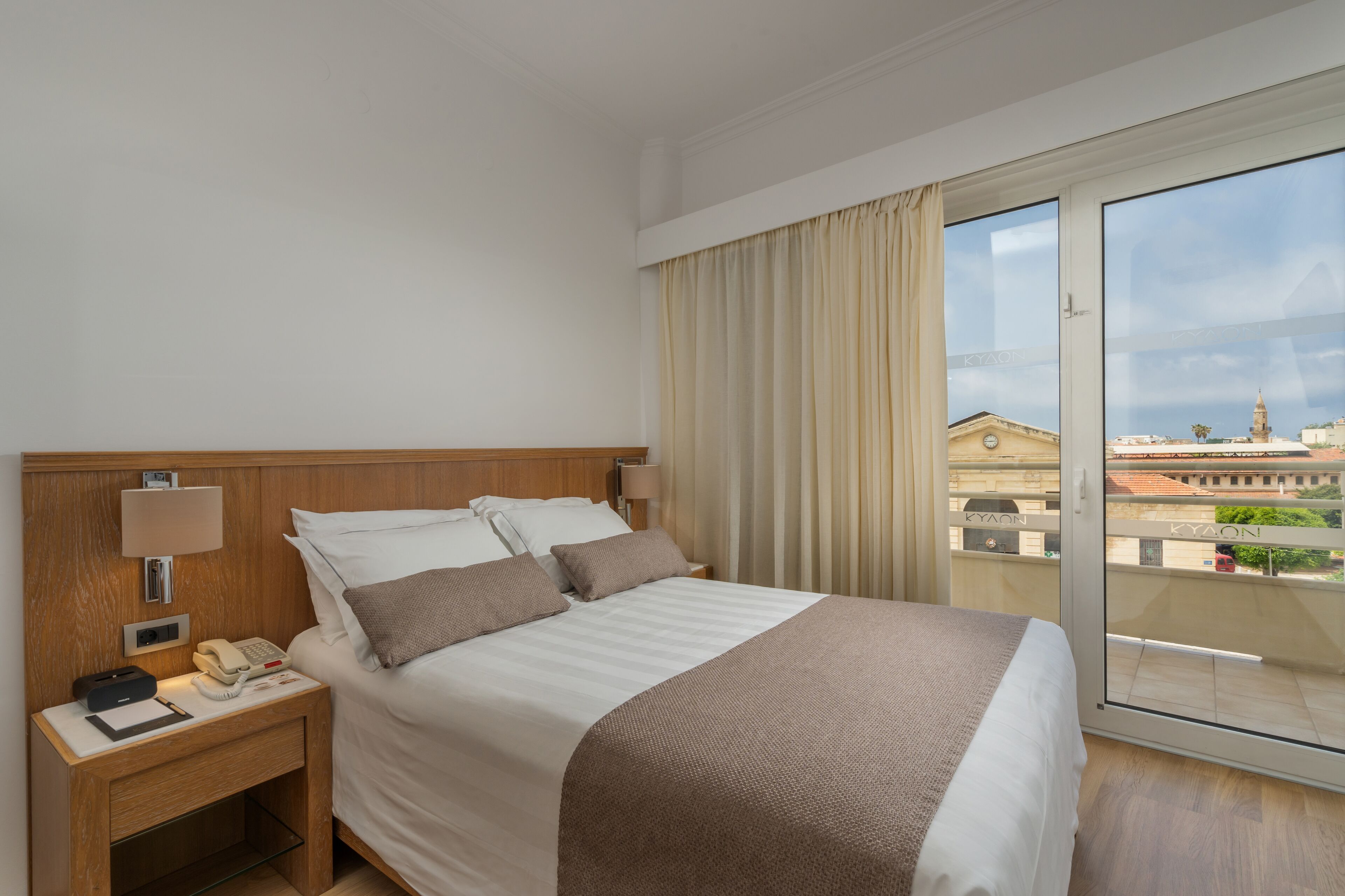 Une chambre d’hôtel avec un grand lit, une table de chevet avec un téléphone et une lampe, et une vue sur la ville depuis un balcon.