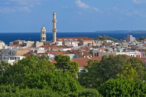 A coastal town with a prominent tower and a mosque, surrounded by buildings with red-tiled roofs and lush greenery in the foreground. - Kydon, The Heart City Hotel (Chania)