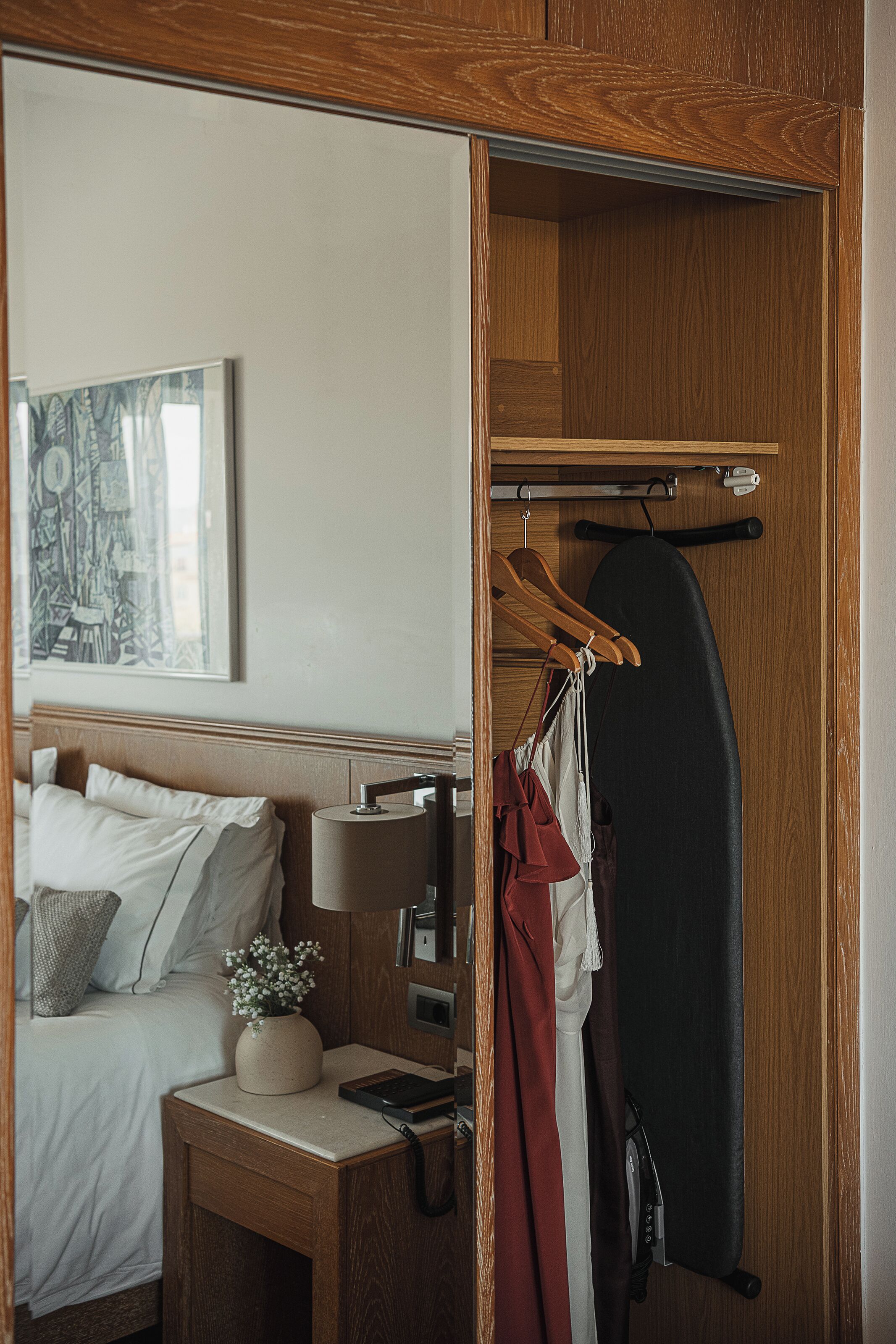 A hotel room with a bed, bedside table, lamp, and a coat rack with hanging clothes.