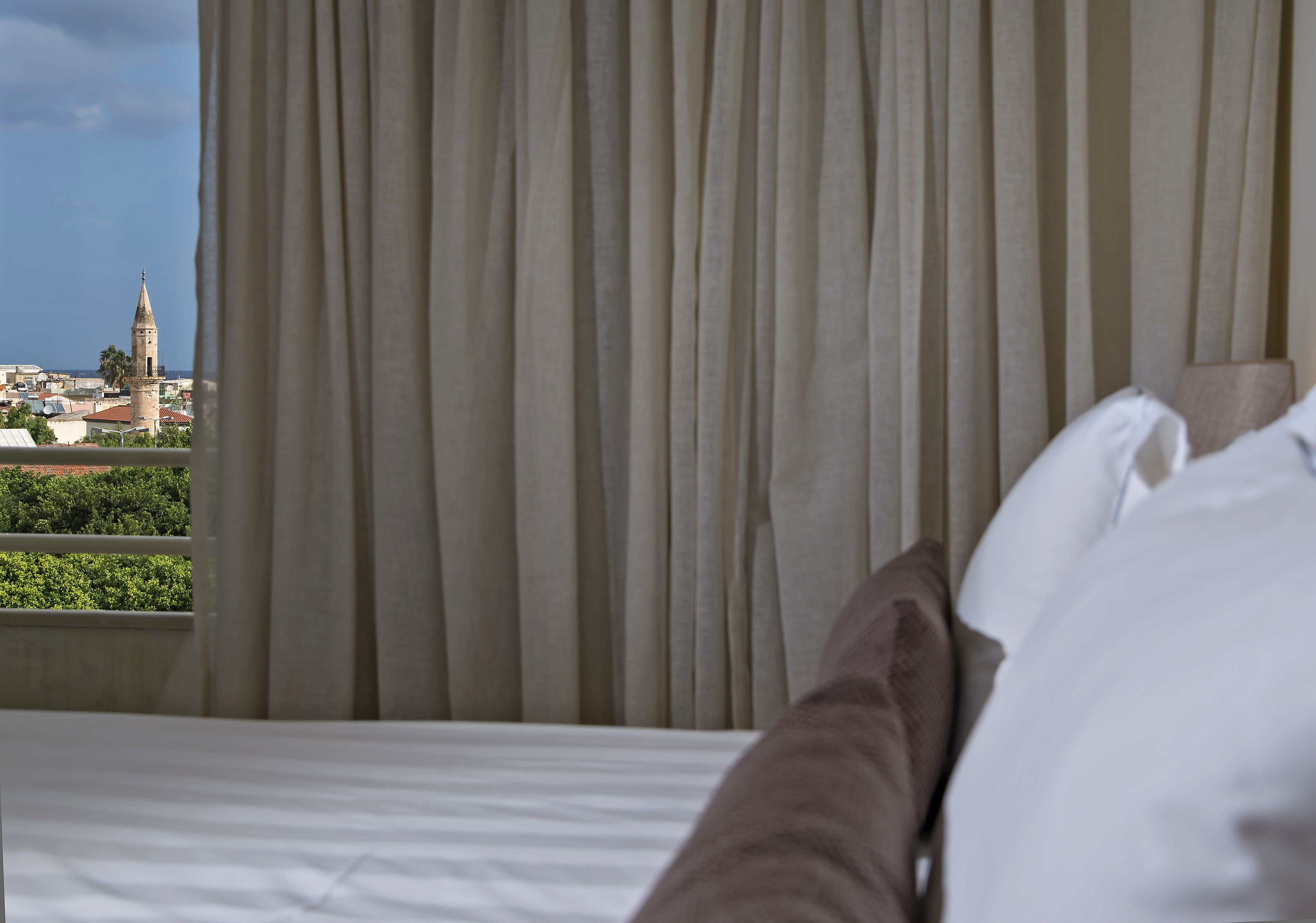 Quarto de hotel com cama, cortinas e vista para uma torre e vegetação.