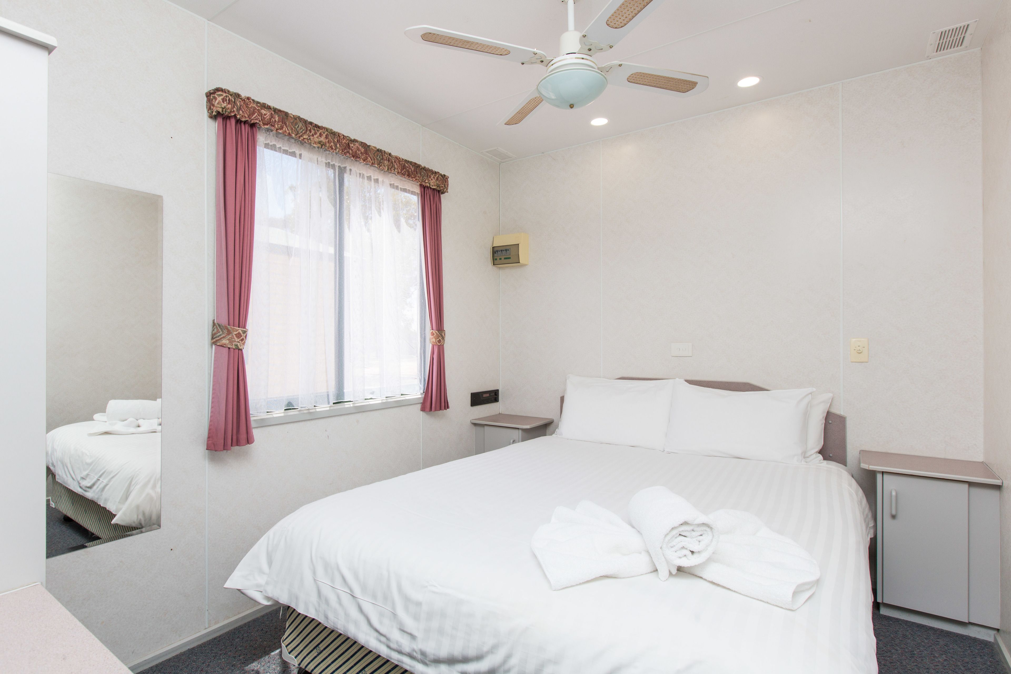 A hotel room with a bed, a nightstand, a mirror, a ceiling fan, and a window with curtains.