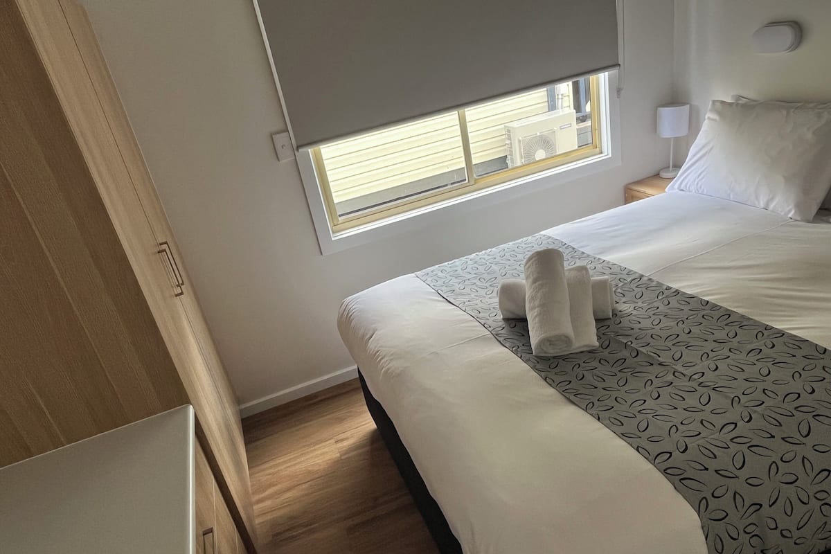 A neatly made bed with white linens and a patterned throw, a window with blinds, and a bedside lamp.