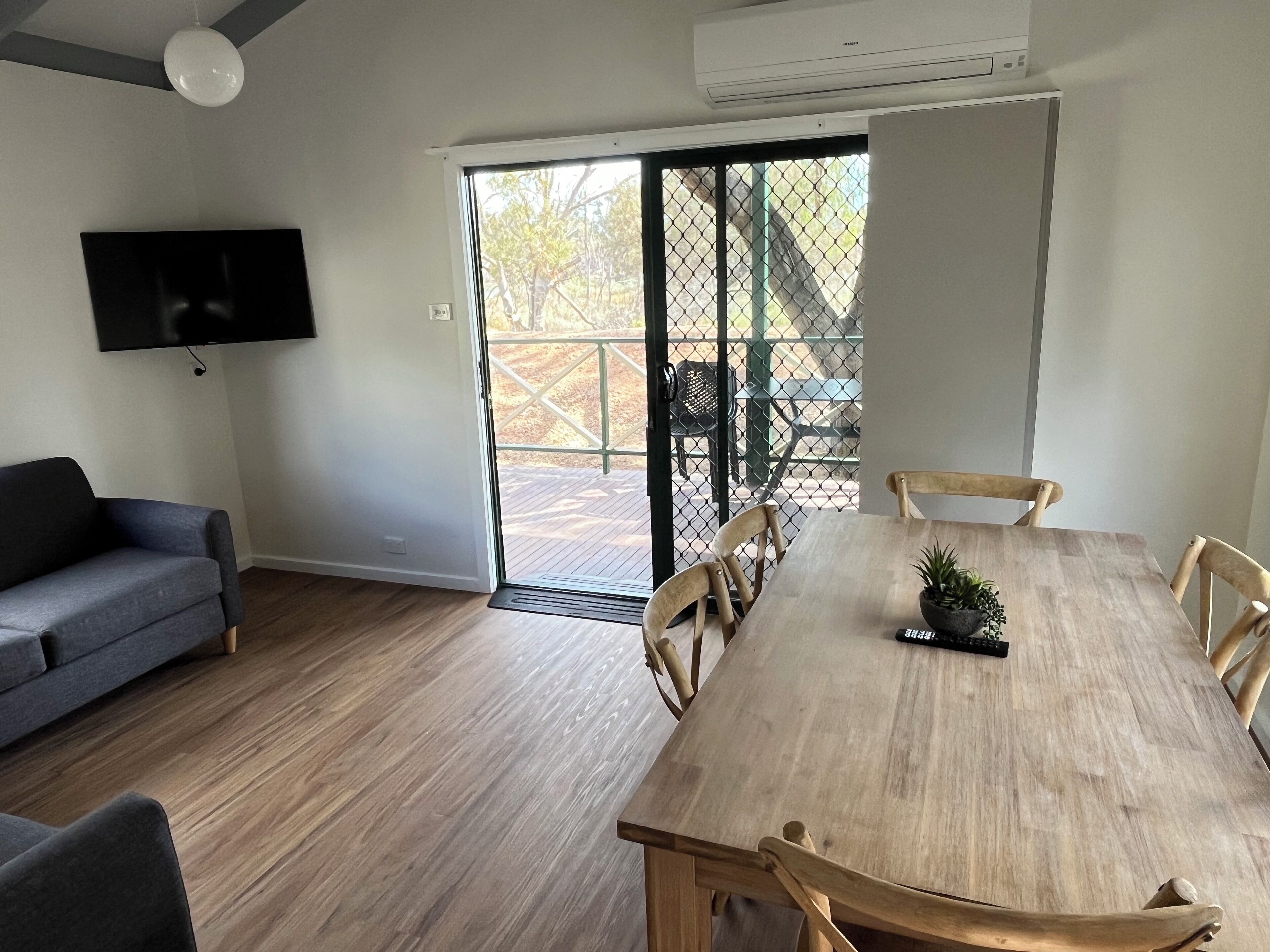 Een moderne woonkamer met een grijze bank, een houten eettafel met stoelen, een aan de muur bevestigde televisie en een schuifdeur van glas die leidt naar een terras.