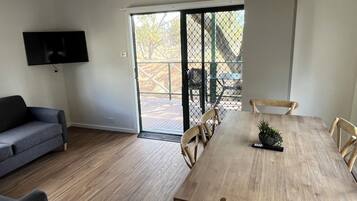 Een moderne woonkamer met een grijze bank, een houten eettafel met stoelen, een aan de muur bevestigde televisie en een schuifdeur van glas die leidt naar een terras.
