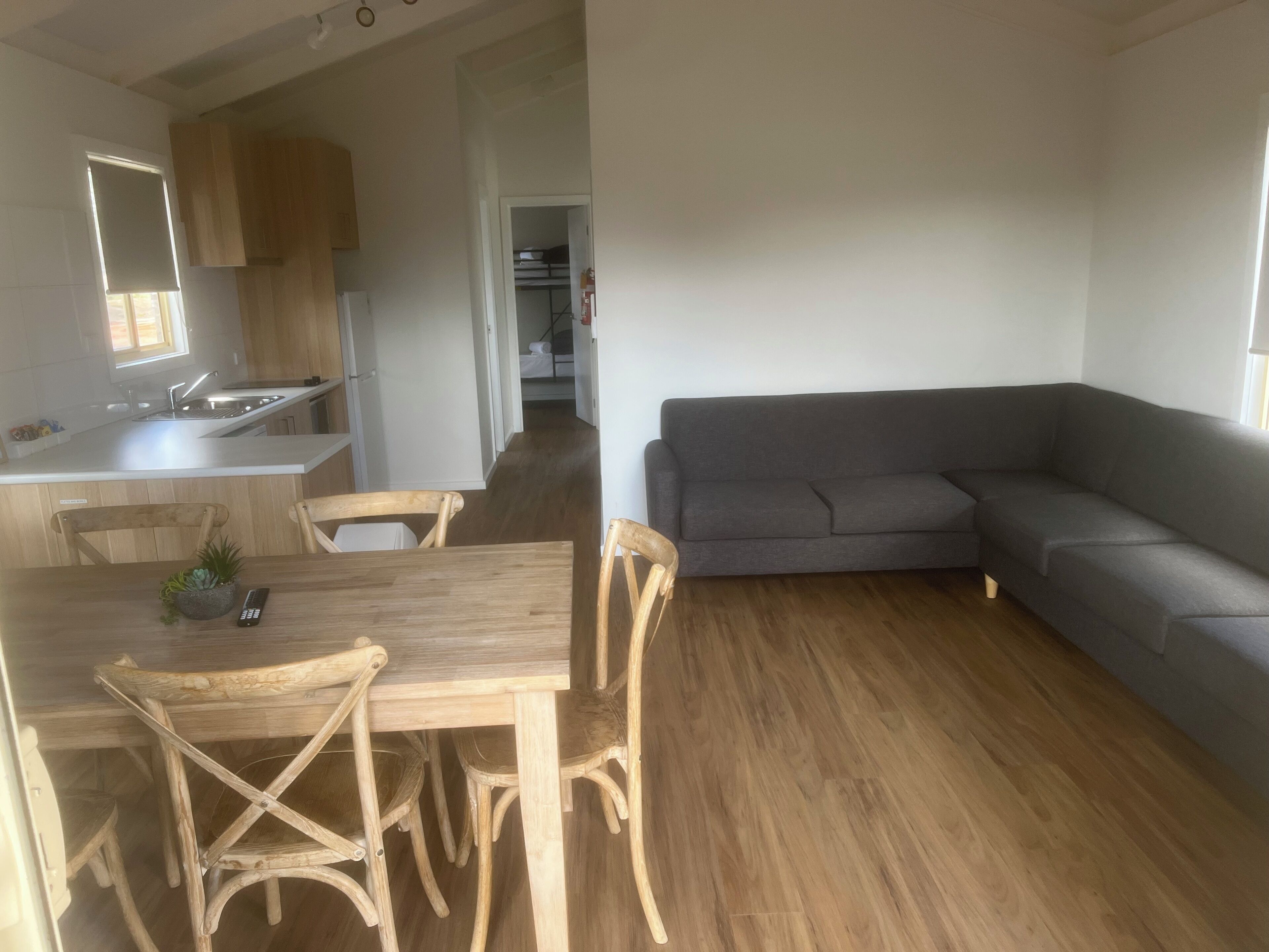 A modern kitchen with wooden cabinets, a dining table with chairs, and a grey sectional sofa in a living area.