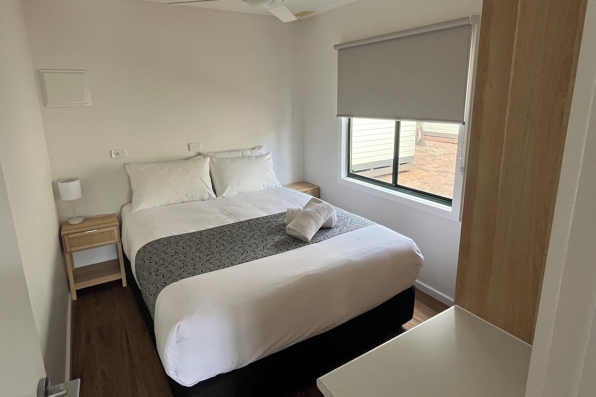 A bedroom with a bed, a nightstand, a ceiling fan, a window with a blind, and a wooden floor.