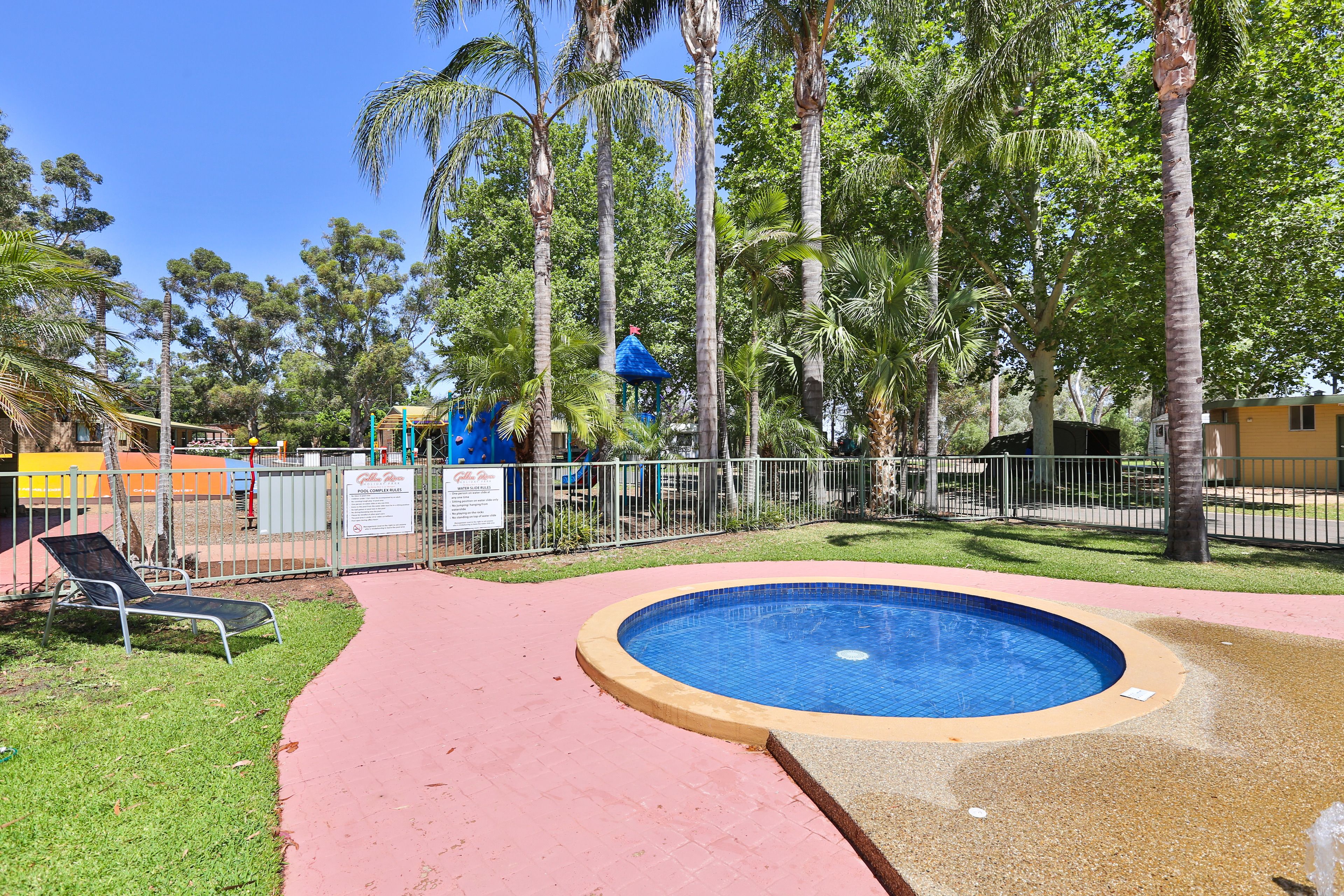 A circular pool with a red walkway, surrounded by palm trees and a fenced area with a blue structure.
