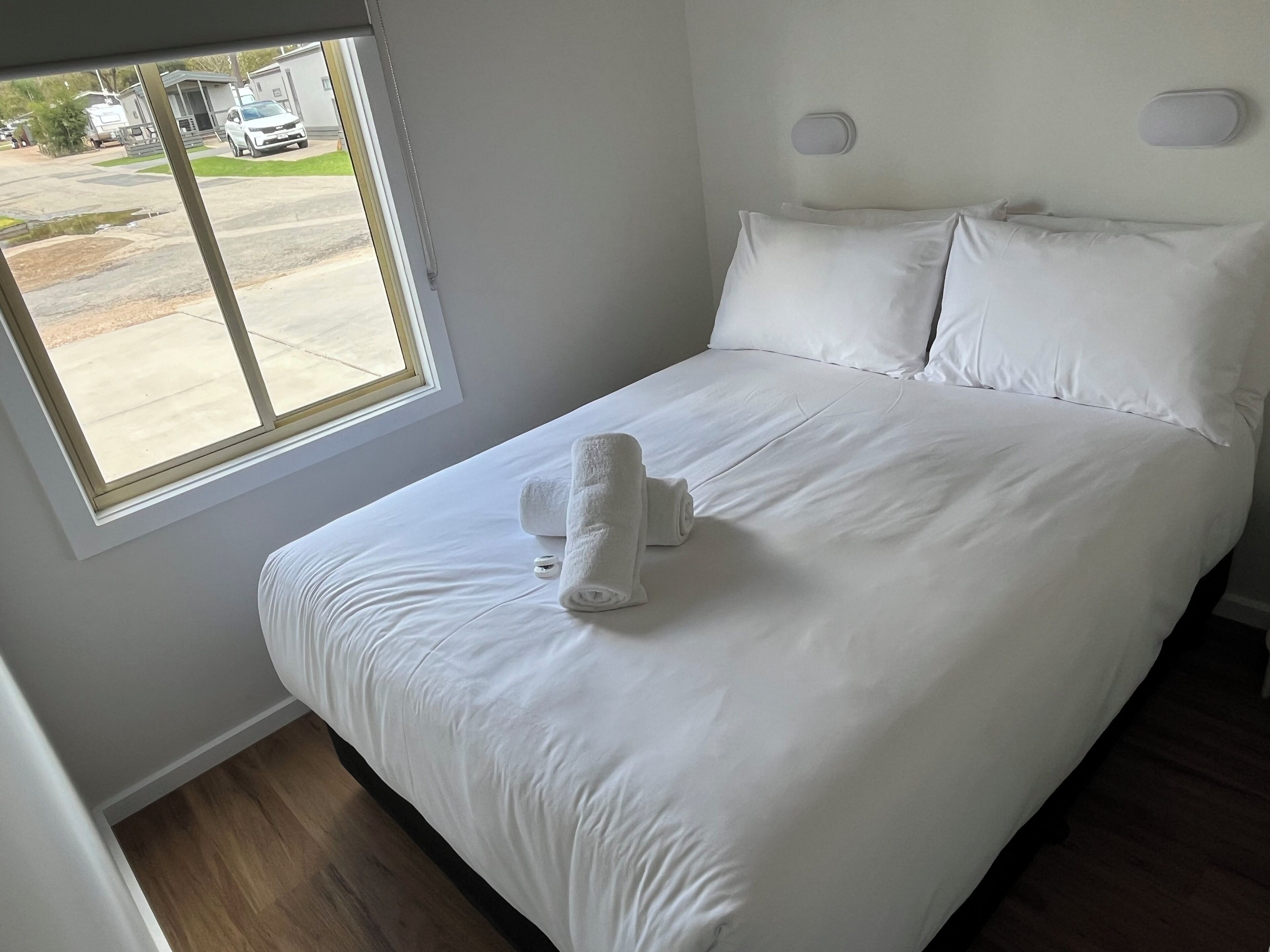 A neatly made bed with white linens and a rolled towel, a window with a view of parked cars and a building outside, and a wall-mounted light fixture.