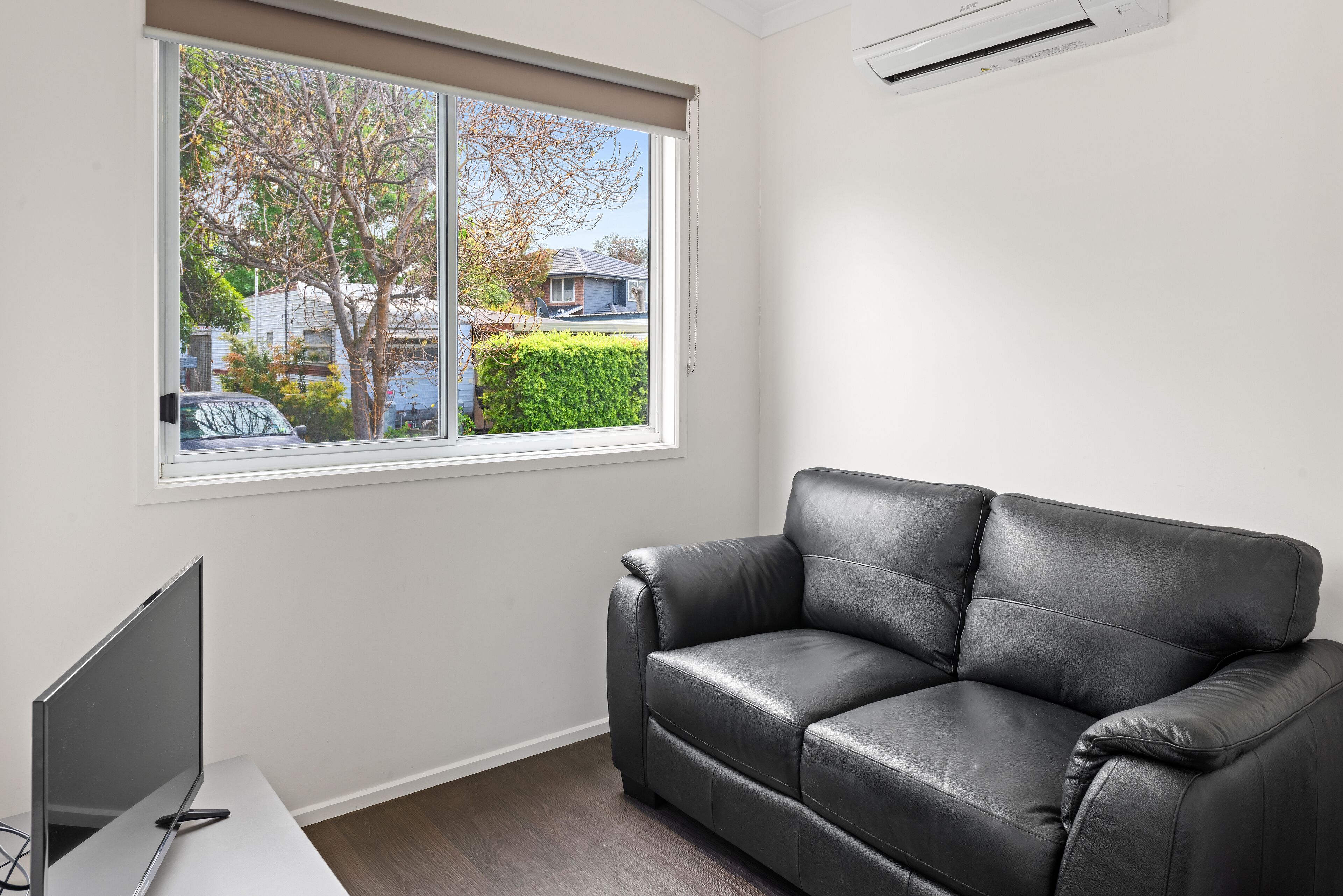 A room with a black leather sofa, a flat-screen TV on a white table, and a window with a view of a residential area.