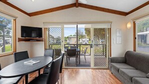 A room with a wooden ceiling, a ceiling fan, a television on a shelf, a dining table with chairs, a sofa, and a sliding glass door leading to an outdoor area. - Discovery Parks - Geelong (Belmont)