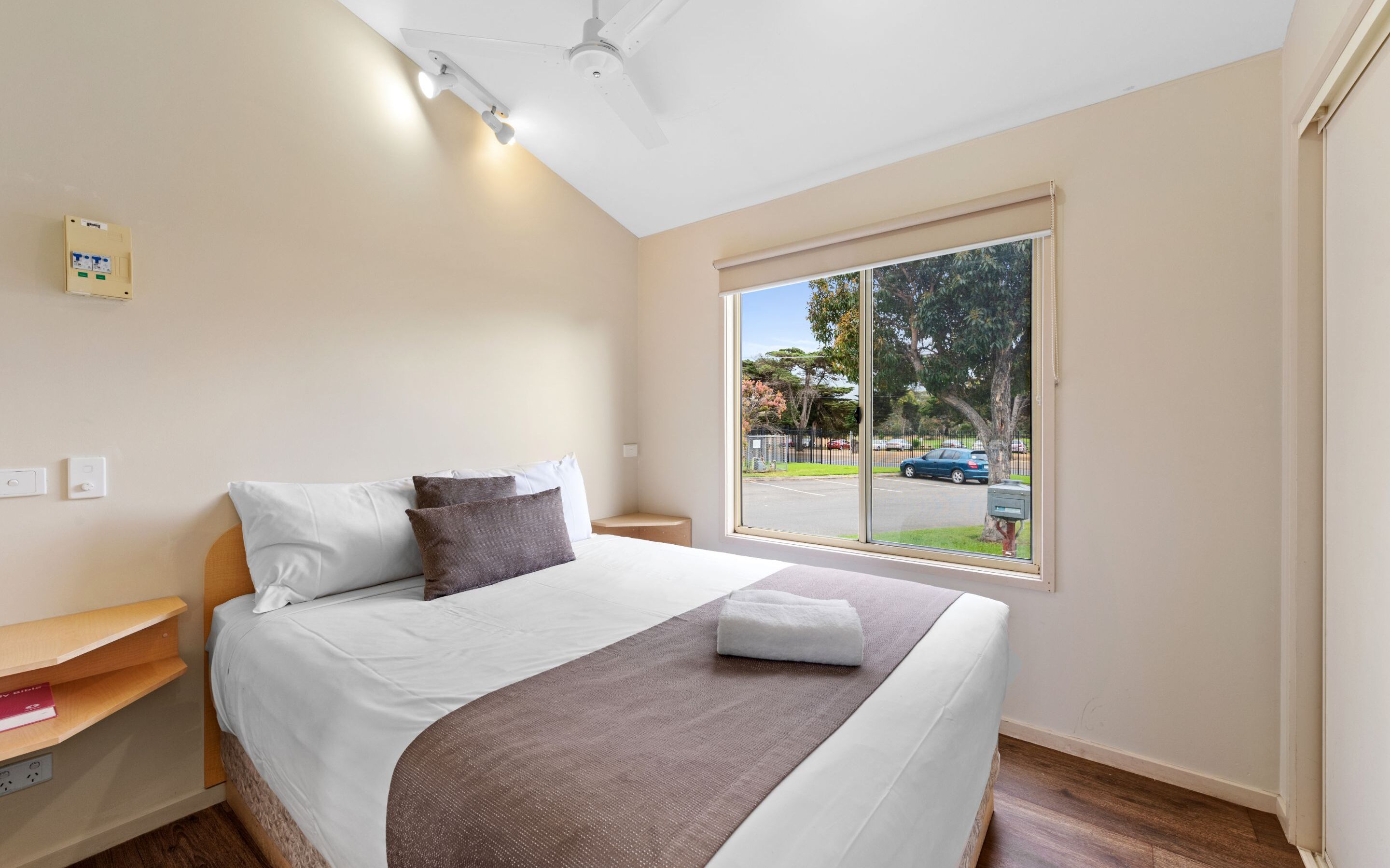 A neatly made bed with white and brown bedding, a bedside table with a book, and a window with a view of the street and cars.