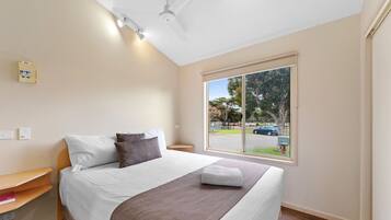 A neatly made bed with white and brown bedding, a bedside table with a book, and a window with a view of the street and cars.