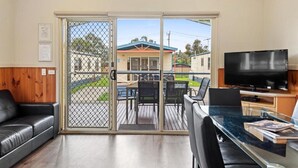 A room with a glass sliding door leading to a deck, a black leather sofa, a glass dining table with chairs, and a flat-screen TV. - Discovery Parks - Geelong (Belmont)