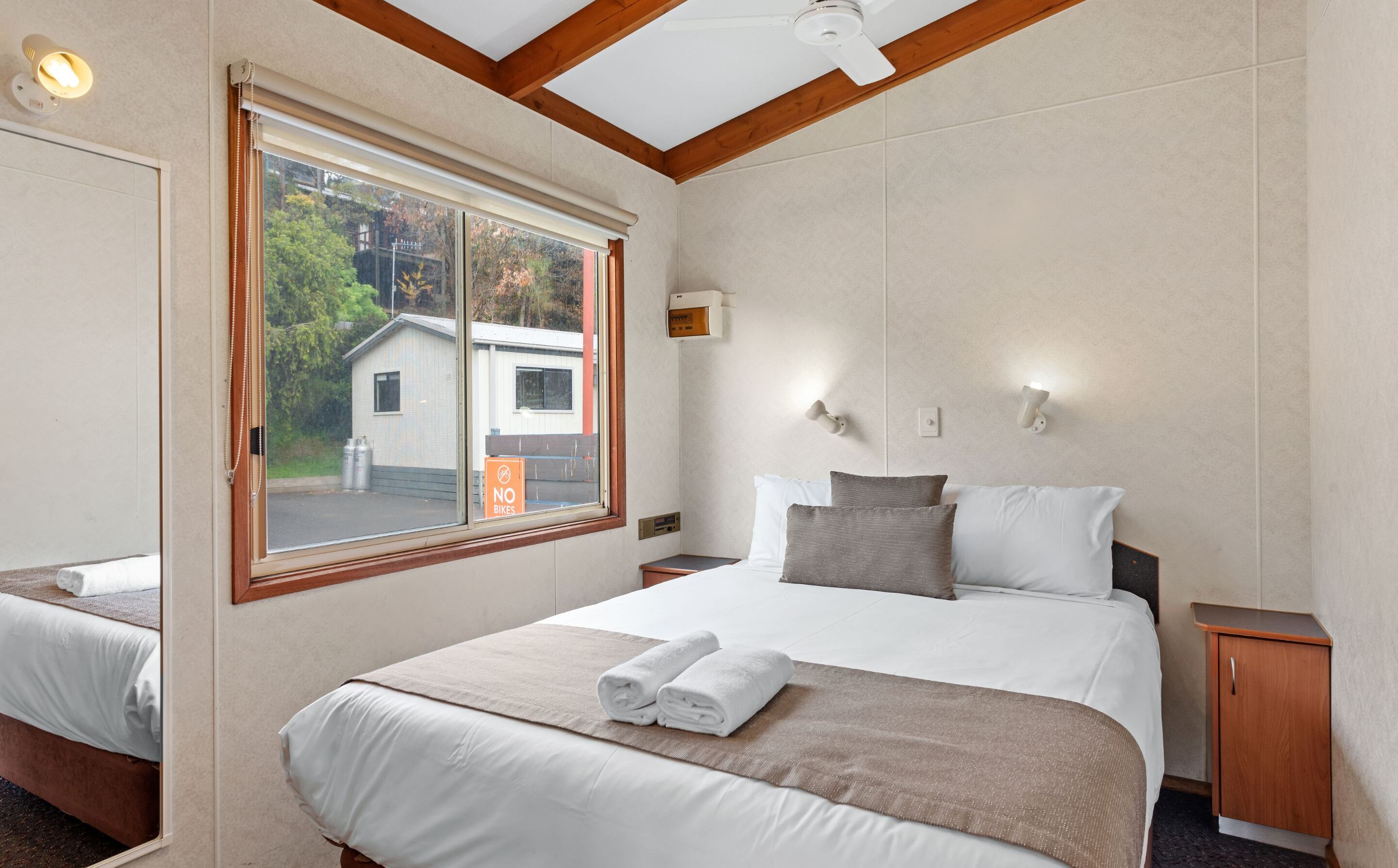 Een hotelkamer met een groot bed, een raam met uitzicht op een gebouw en bomen, een plafondventilator en een nachtkastje.