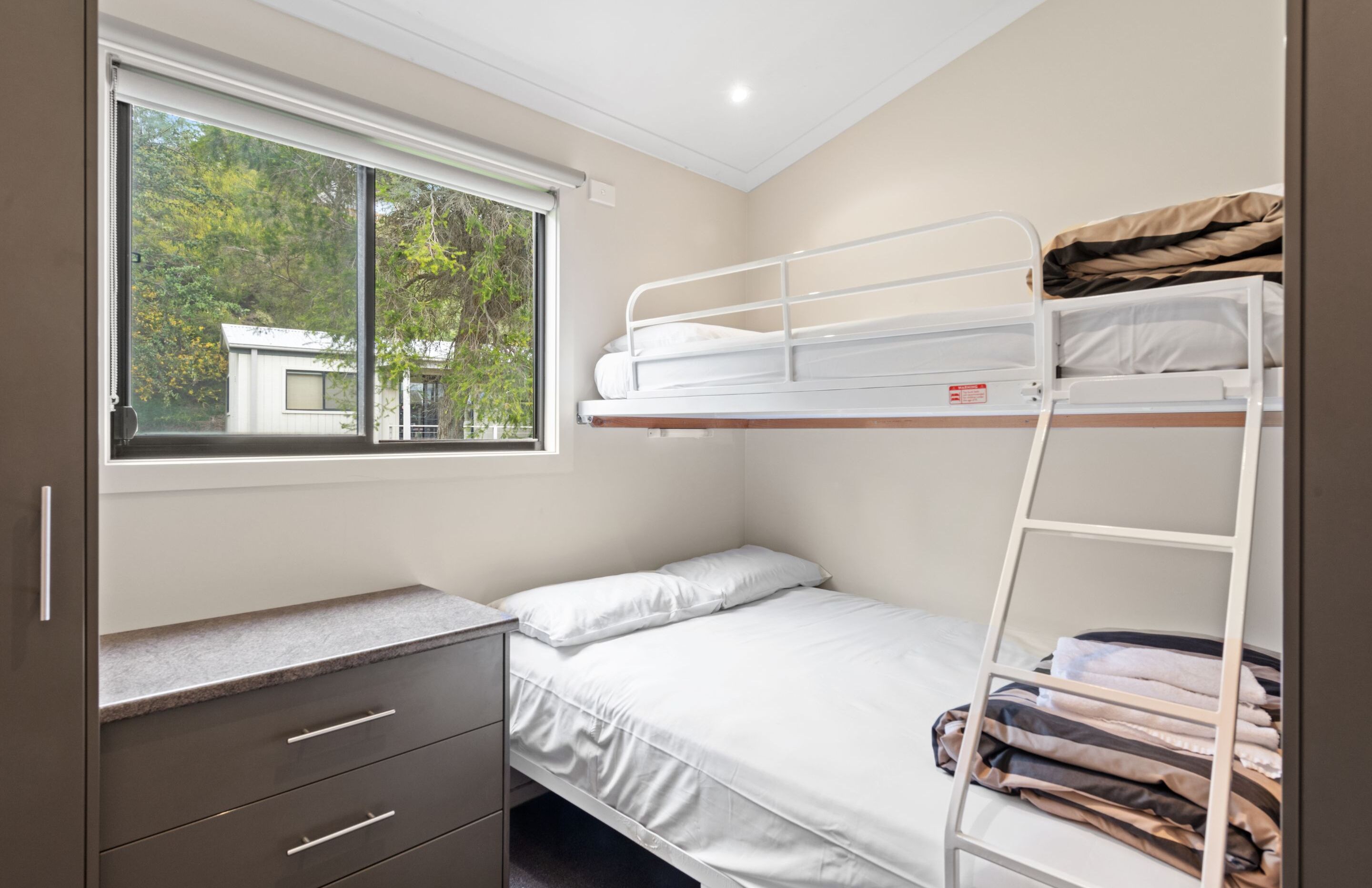 A bunk bed room with a window, a dresser, and a view of trees and a building outside.