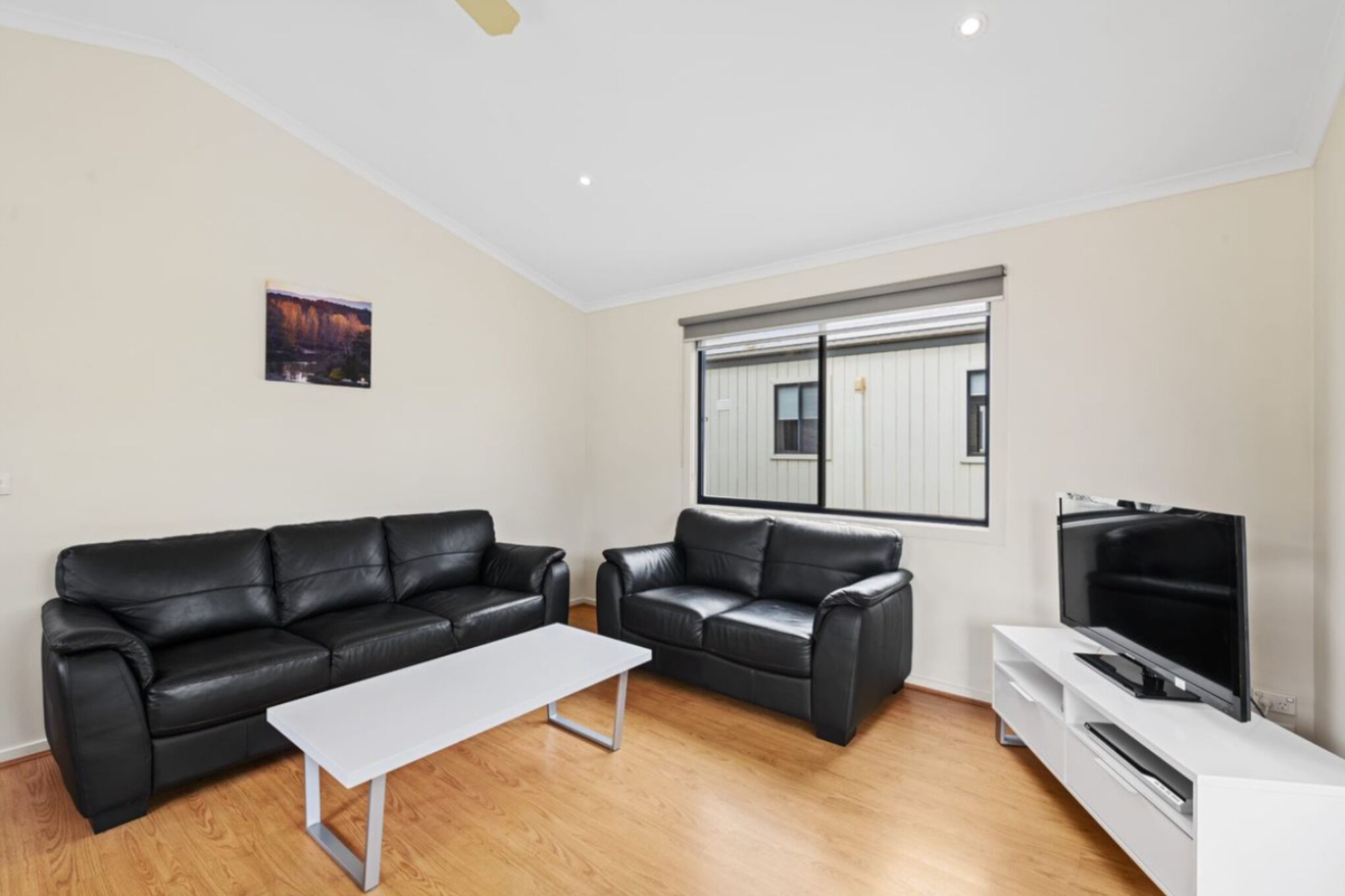 Una sala de estar moderna con dos sofás de cuero negro, una mesa de centro blanca, un televisor de pantalla plana sobre un soporte blanco y una ventana con persianas.