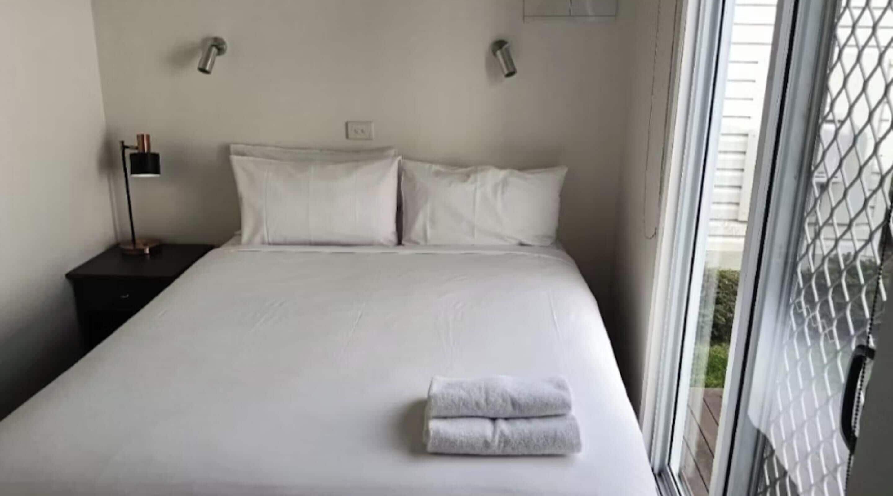 Un lit bien fait, avec du linge de lit blanc et des oreillers, une petite table de chevet avec une lampe, et une fenêtre donnant sur un paysage verdoyant.