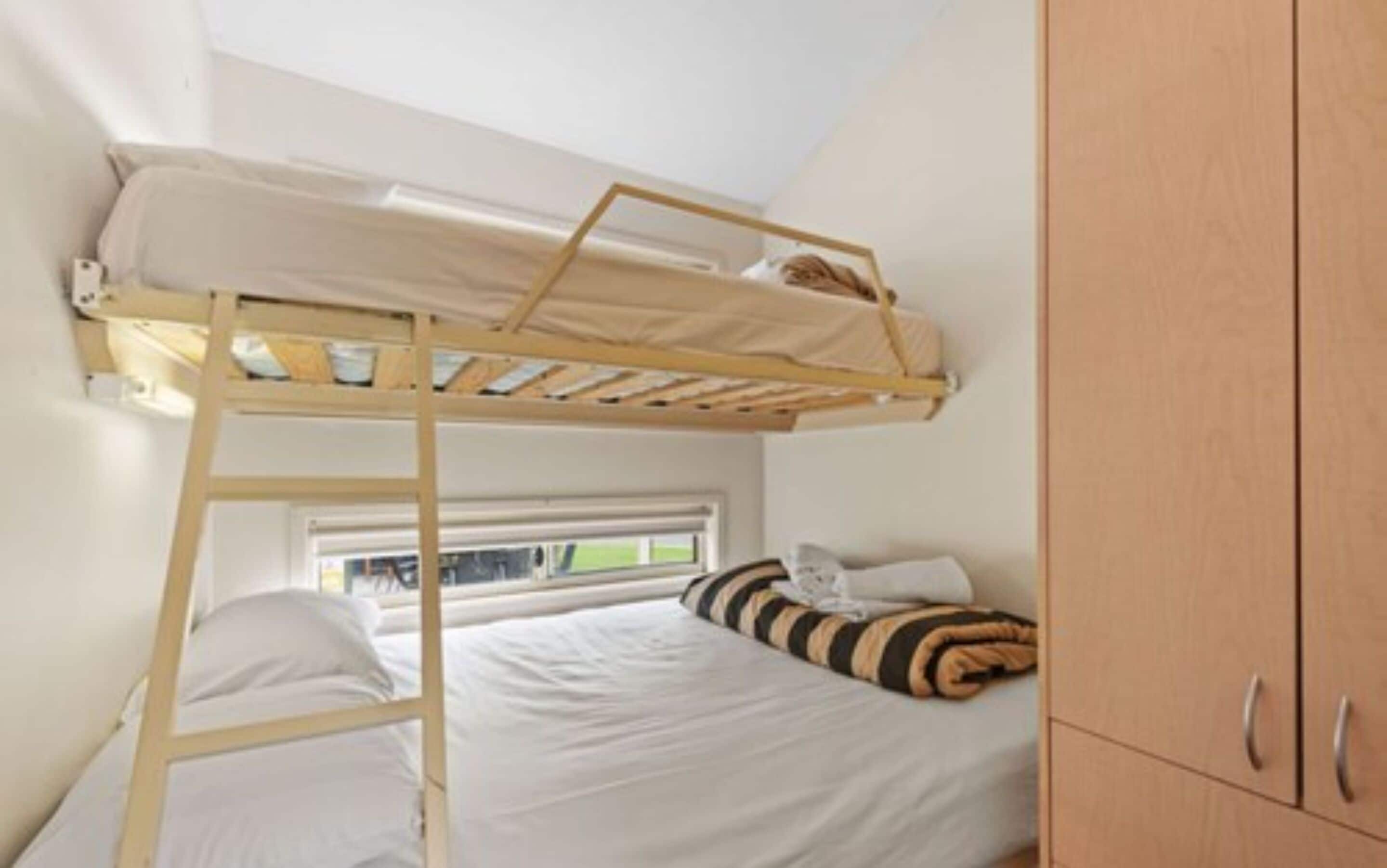 Una cama litera con escalera de madera, una ventana con persianas y un armario de madera.