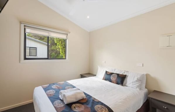 A neatly arranged bedroom with a bed, a bedside table, a window with blinds, and a view of a building outside. - Discovery Parks - Geelong (Belmont)