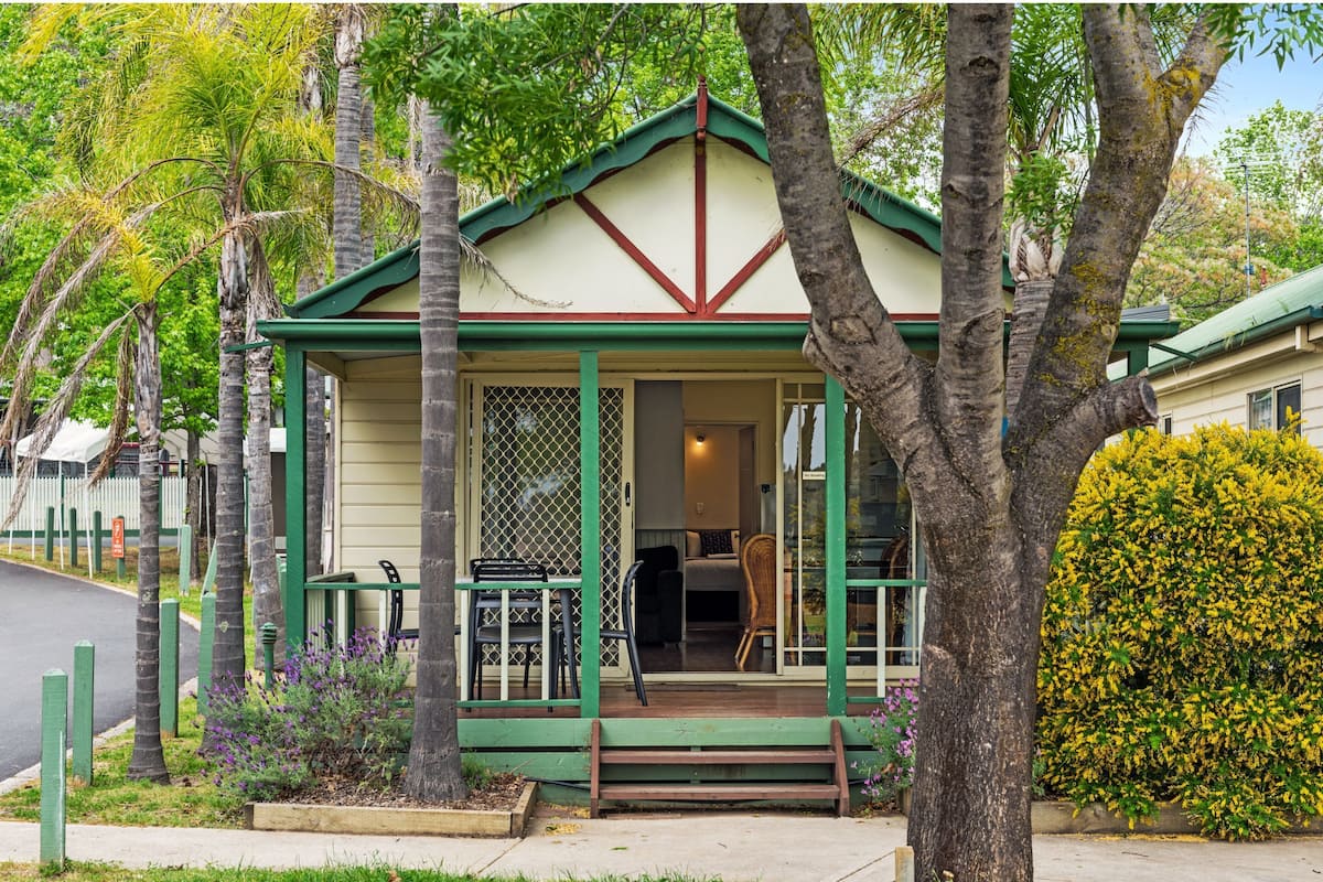 A small, single-story building with a green roof and white walls, featuring a porch with outdoor seating and a visible interior with a bed and a chair.