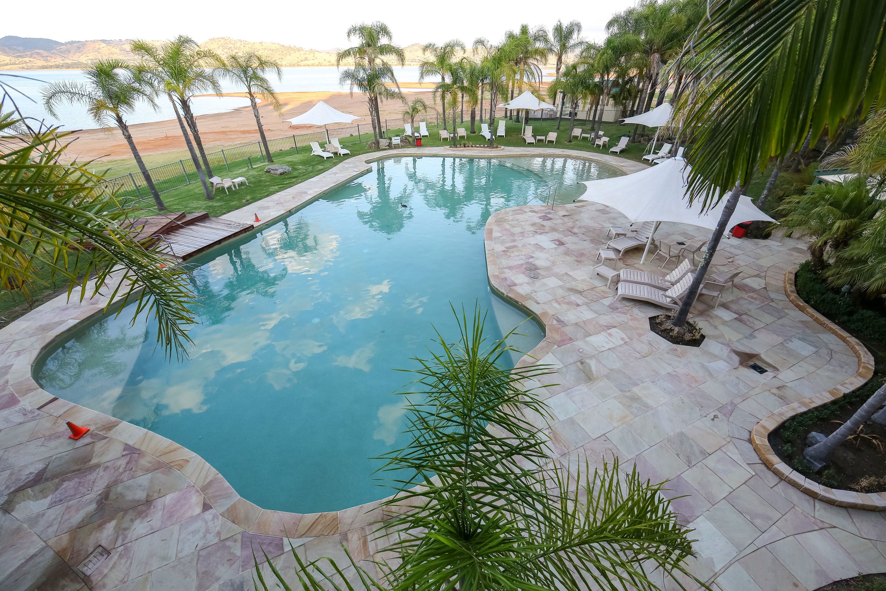 Una piscina de borde curvo, rodeada de sillones de baño y sombrillas, con vista a una playa y montañas al fondo.