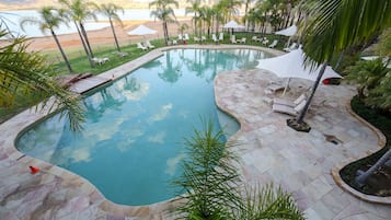 Una piscina de borde curvo, rodeada de sillones de baño y sombrillas, con vista a una playa y montañas al fondo.