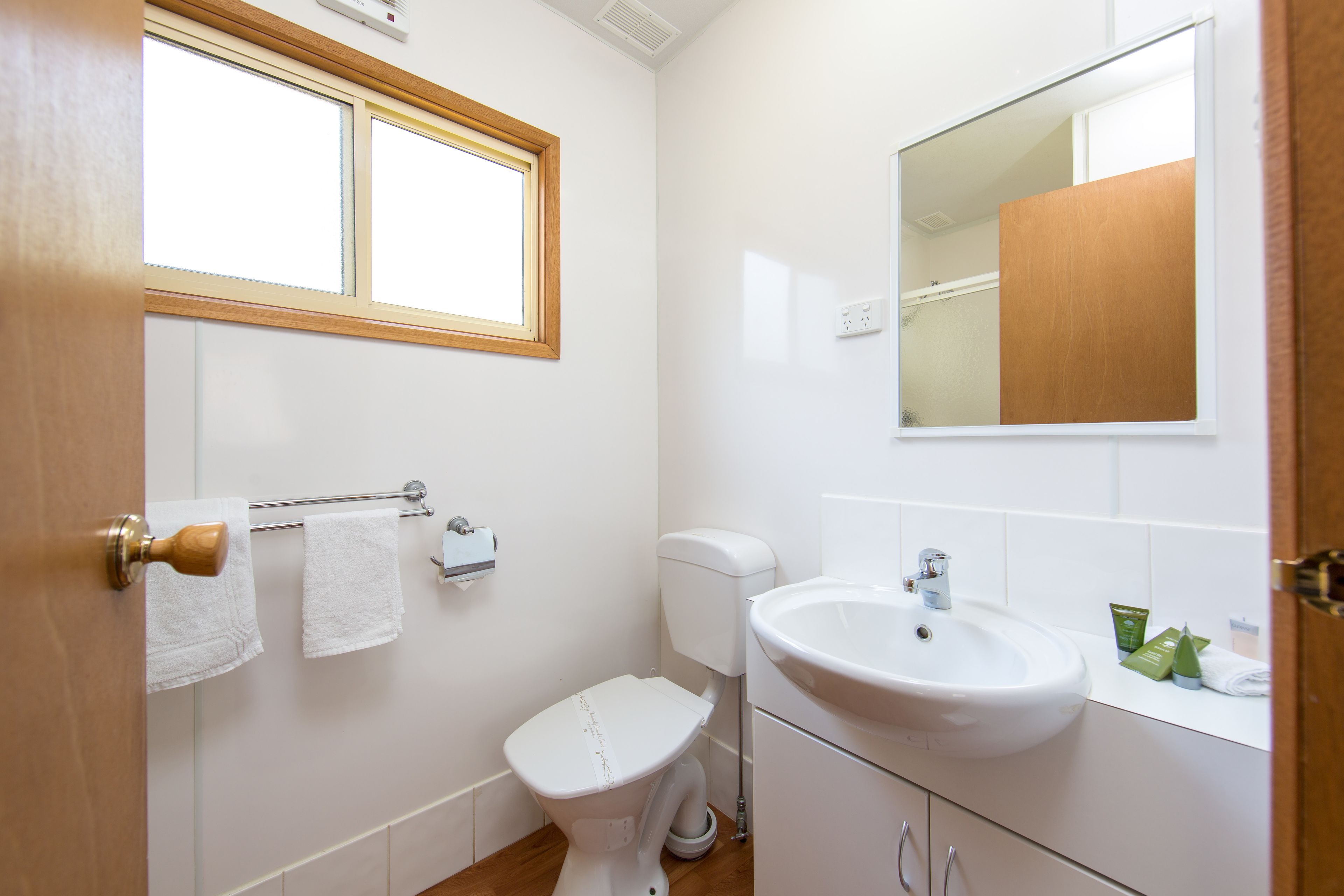 A bathroom with a toilet, sink, and mirror. There are towels hanging on a rack and a window above the toilet.