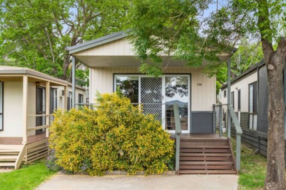 A single-story mobile home with a front porch, a small front yard, and a tree to the right.