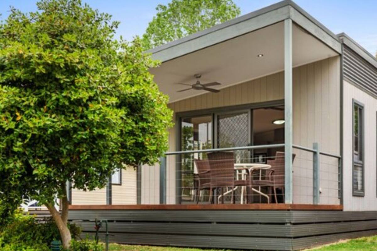 A modern single-story house with a balcony, large windows, and a tree in the foreground.