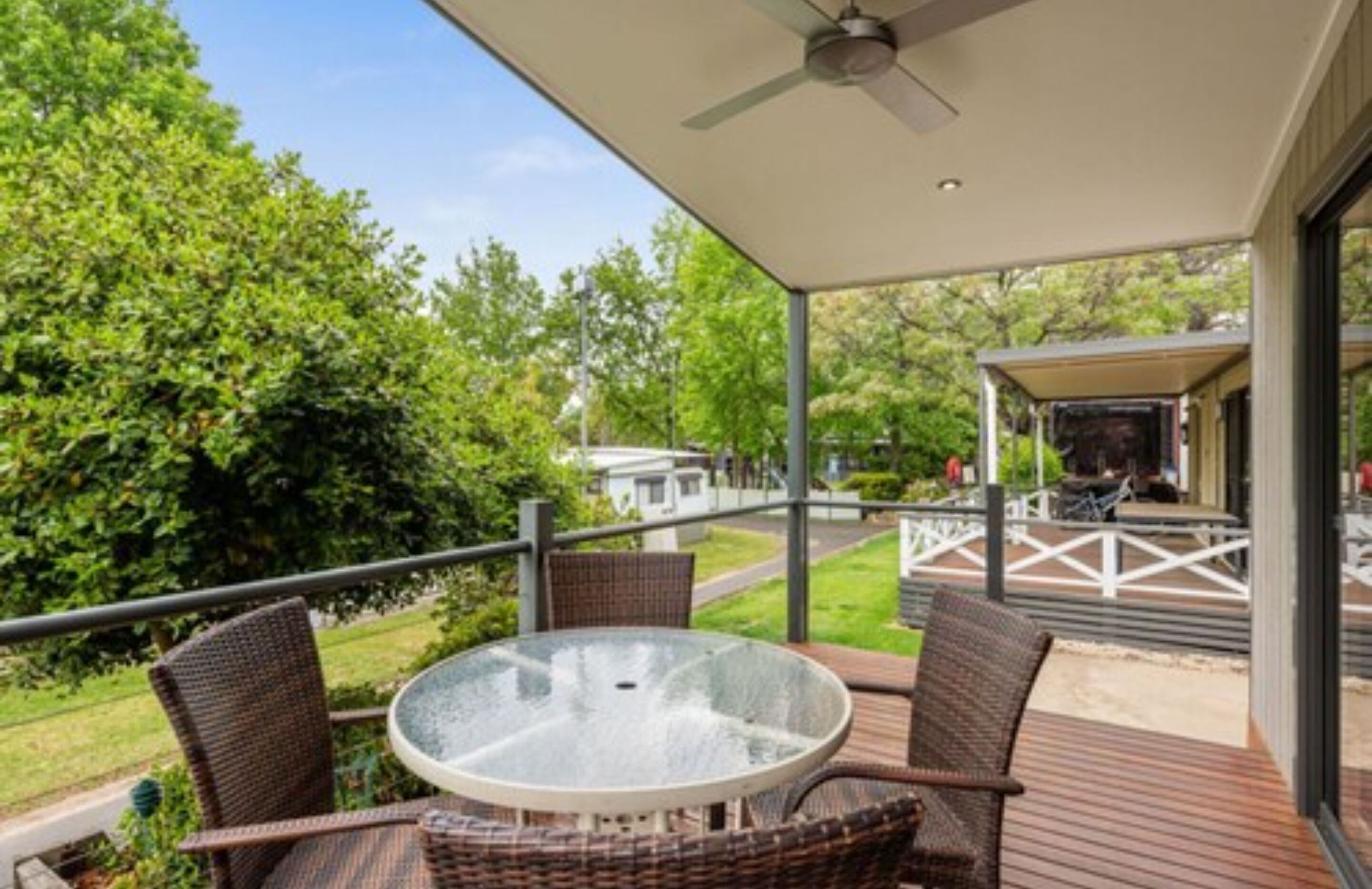 A covered outdoor area with a glass table and wicker chairs, overlooking a well-maintained lawn and a building in the background.