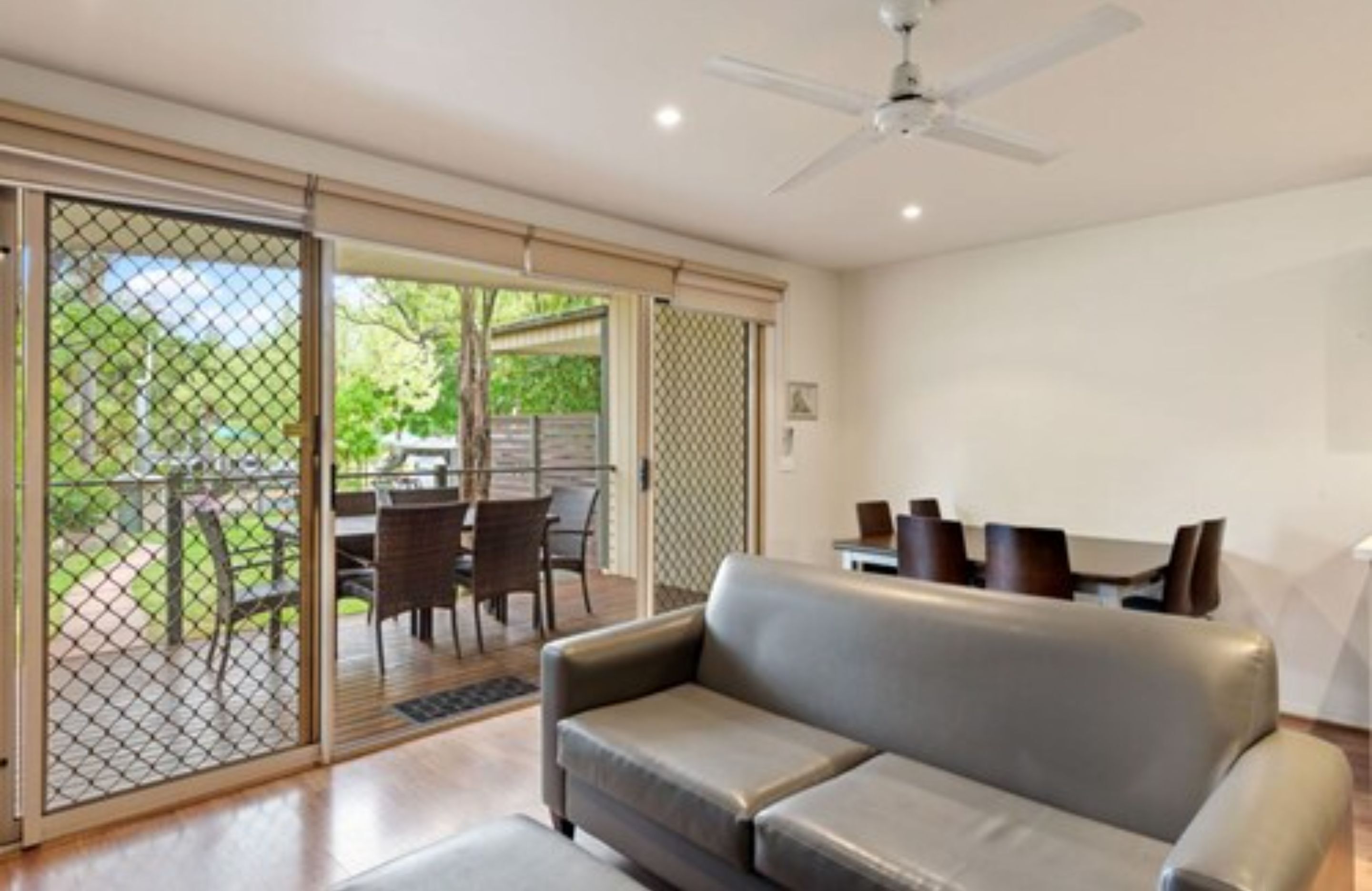 A modern living room with a grey sofa, a ceiling fan, and a sliding glass door leading to an outdoor dining area.