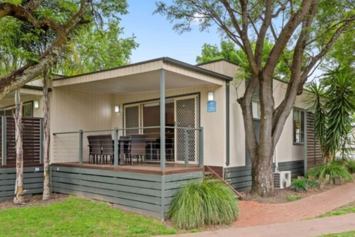 A single-story mobile home with a covered porch, a dining area, and a small garden area with trees and shrubs.