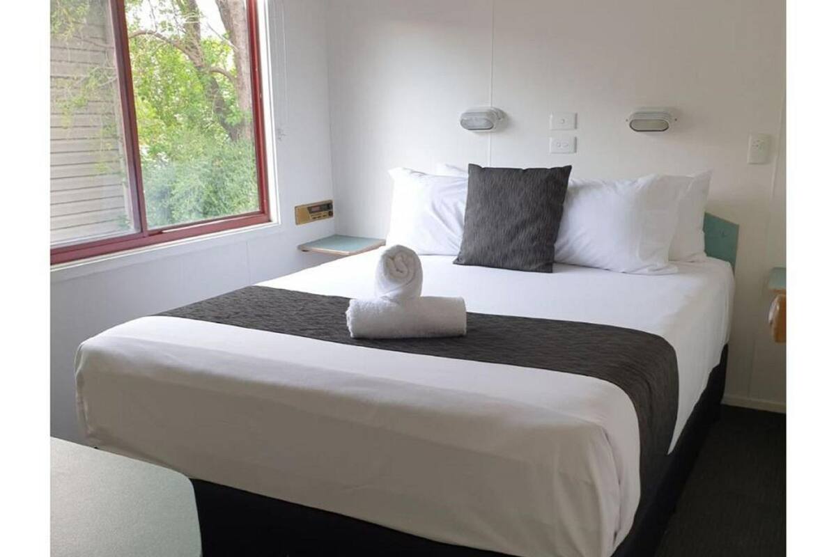 Una cama bien hecha con sábanas blancas y una toalla doblada, una mesita de noche con una lámpara, y una ventana con vistas a árboles.
