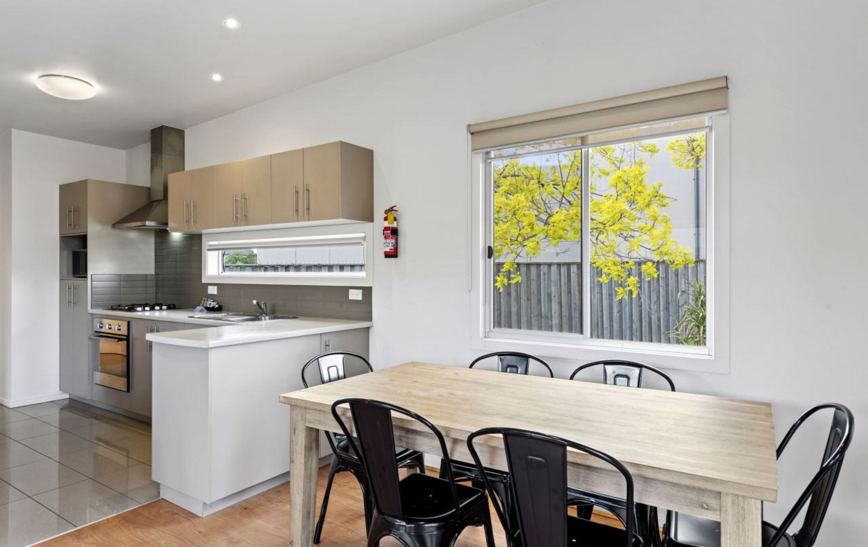 A modern kitchen with a central island, a dining table with chairs, and a window with a view of trees.
